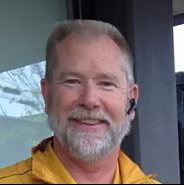Smiling person in a yellow jacket standing by a window, wearing an earbud.