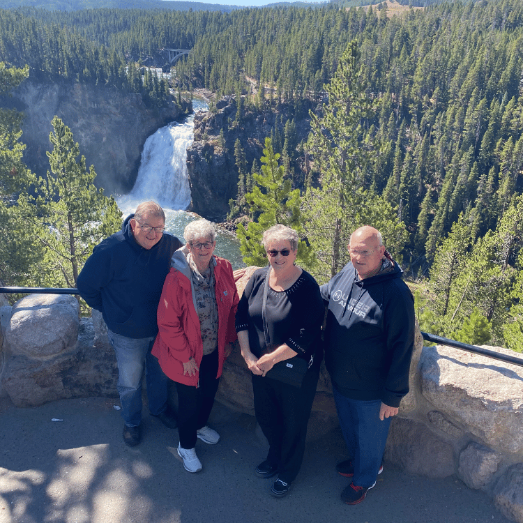 Four people pose in front of a waterfall and forest; sunny day, stone ledge.