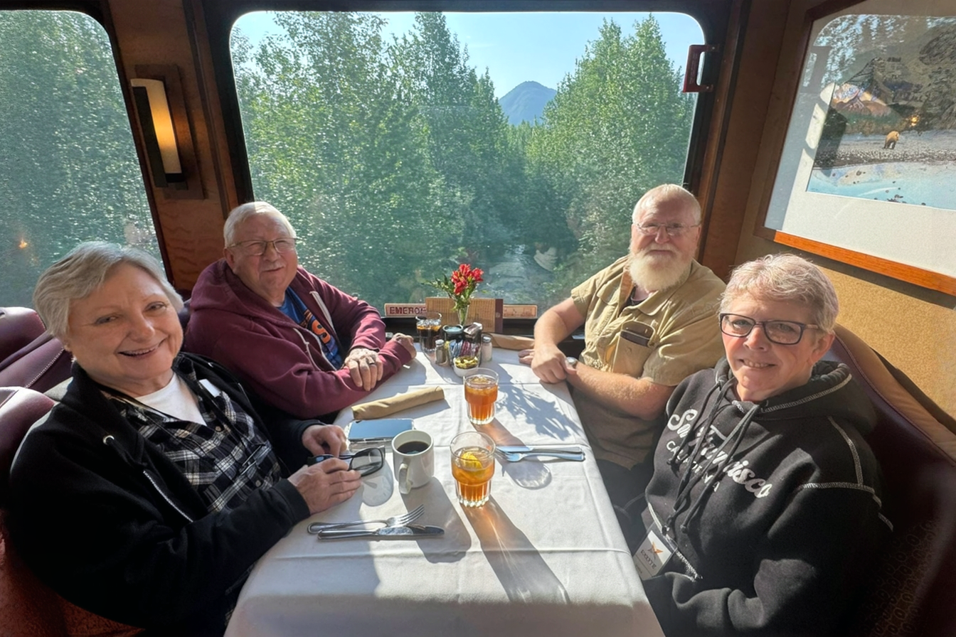 Four people dine together inside a train car, looking out at passing trees and water.