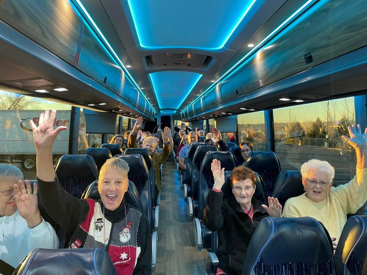 People waving from inside a bus with blue overhead lights. Outside is a sunny landscape.