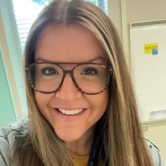 Woman with glasses smiling, long blonde hair, inside with whiteboard and blinds in the background.