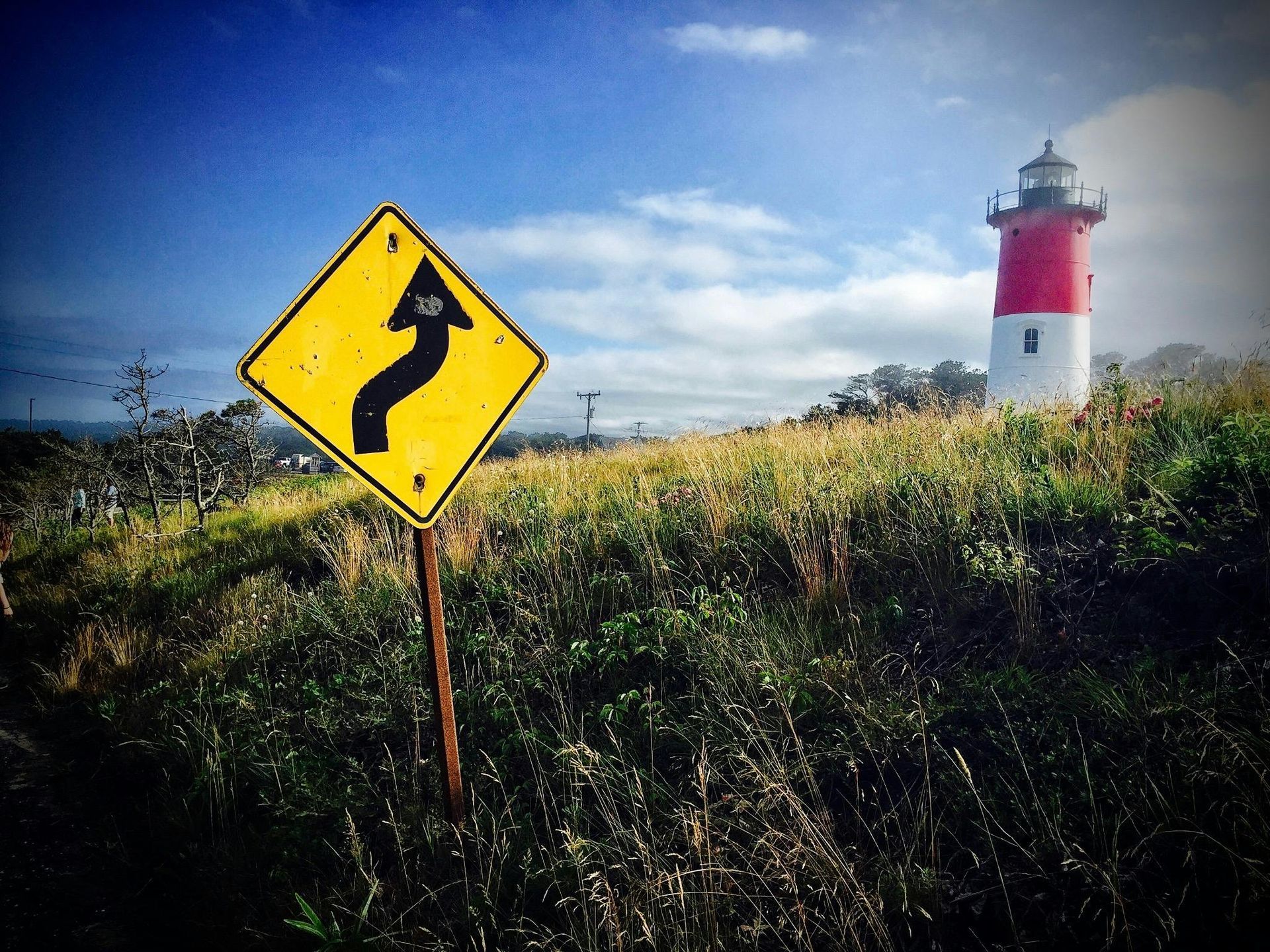 Yellow road sign for curves on a grassy hill, with a red and white lighthouse under a blue sky.