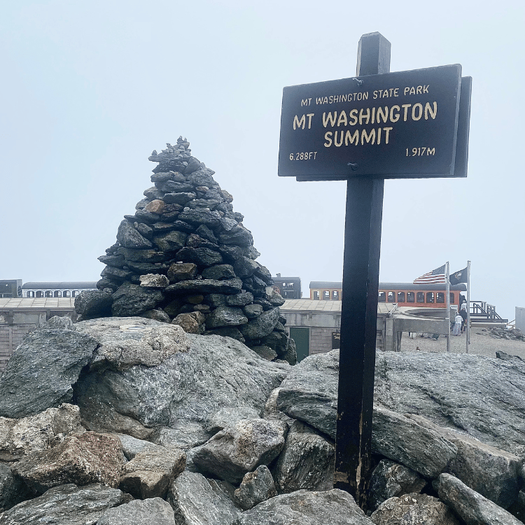 Sign at Mt. Washington Summit with a cairn, foggy. Building, flags visible in background.