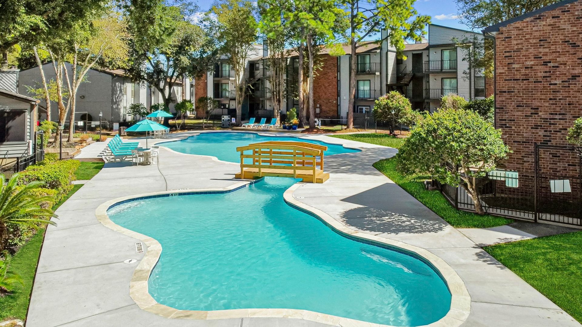 Outdoor apartment pool with loungers, umbrellas, a wooden bridge, and surrounding trees.