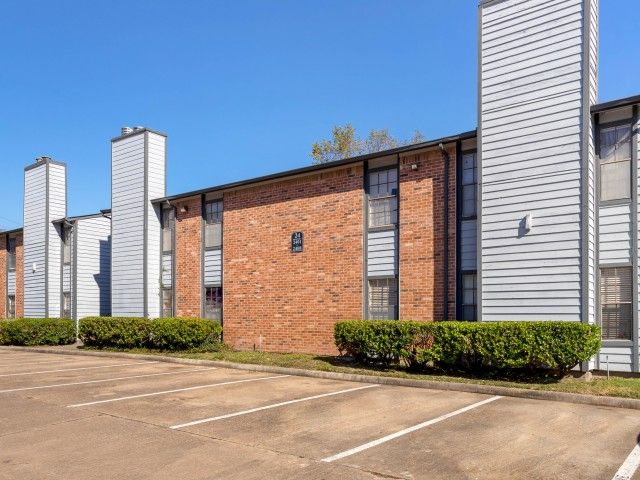 Exterior view of a brick apartment building with a parking lot and trimmed hedges under a clear blue sky.