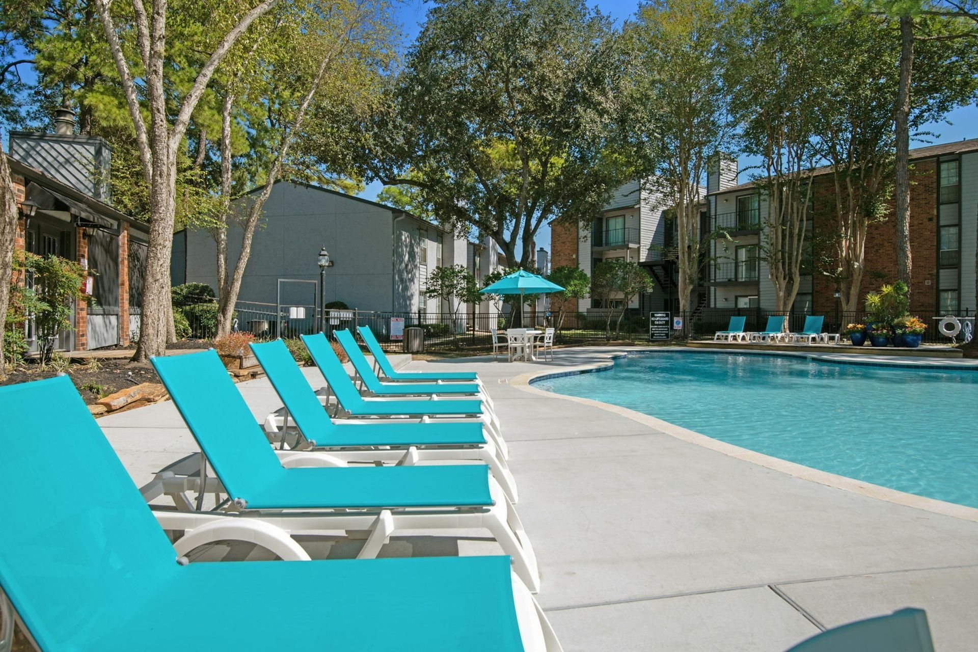 Outdoor apartment community pool with turquoise lounge chairs and trees.