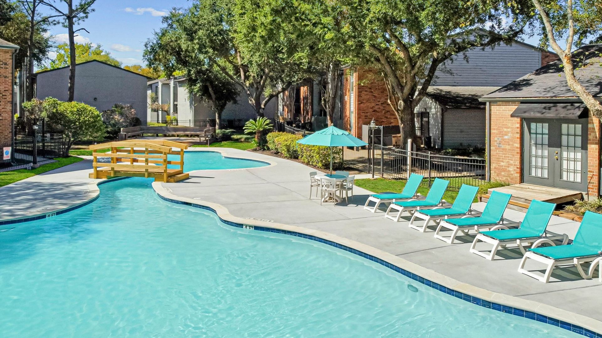 Outdoor community pool with turquoise water, a wooden bridge, and lounge chairs.