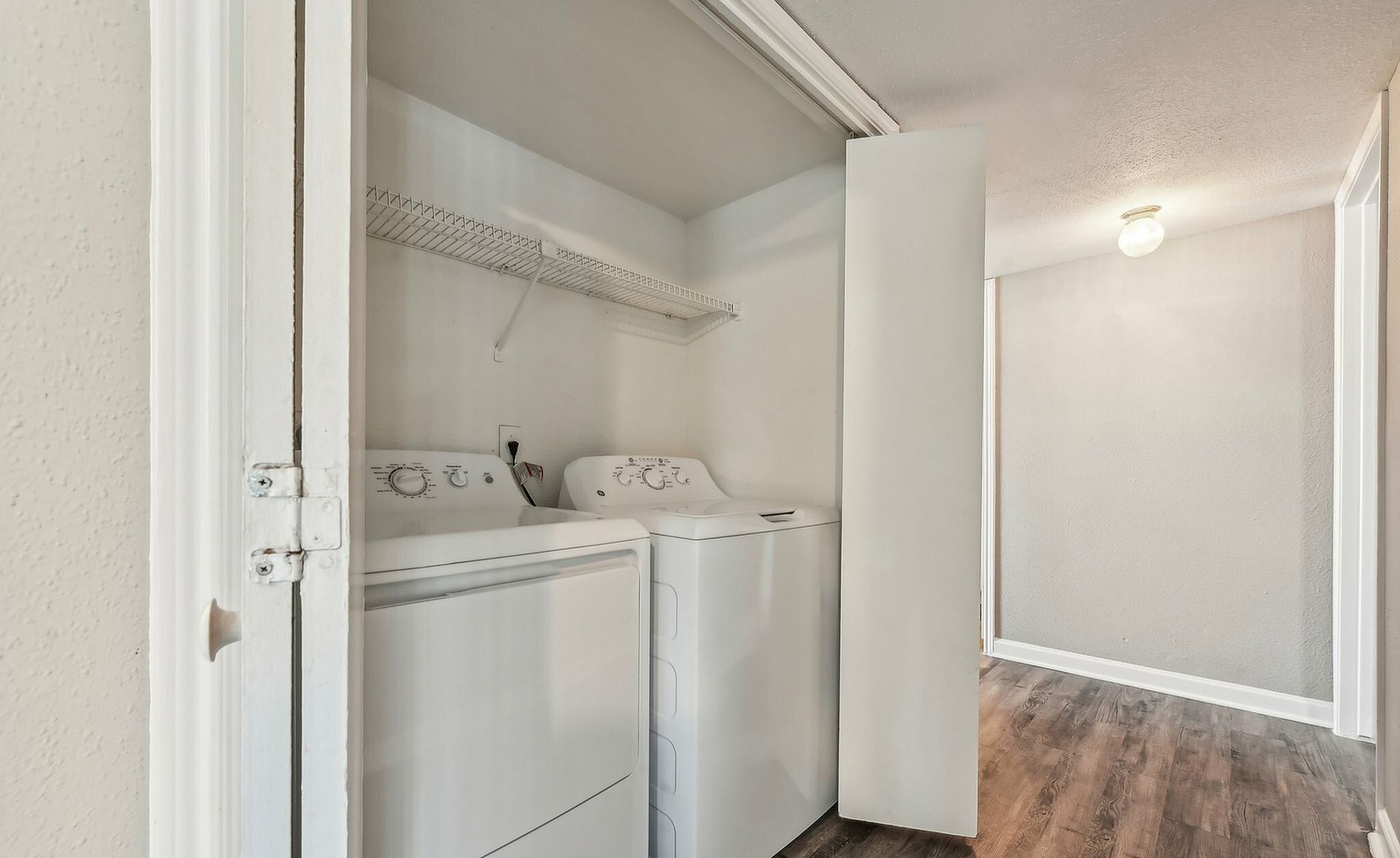 Compact laundry closet with washer and dryer, shelf above, opening to a wood-floor room.