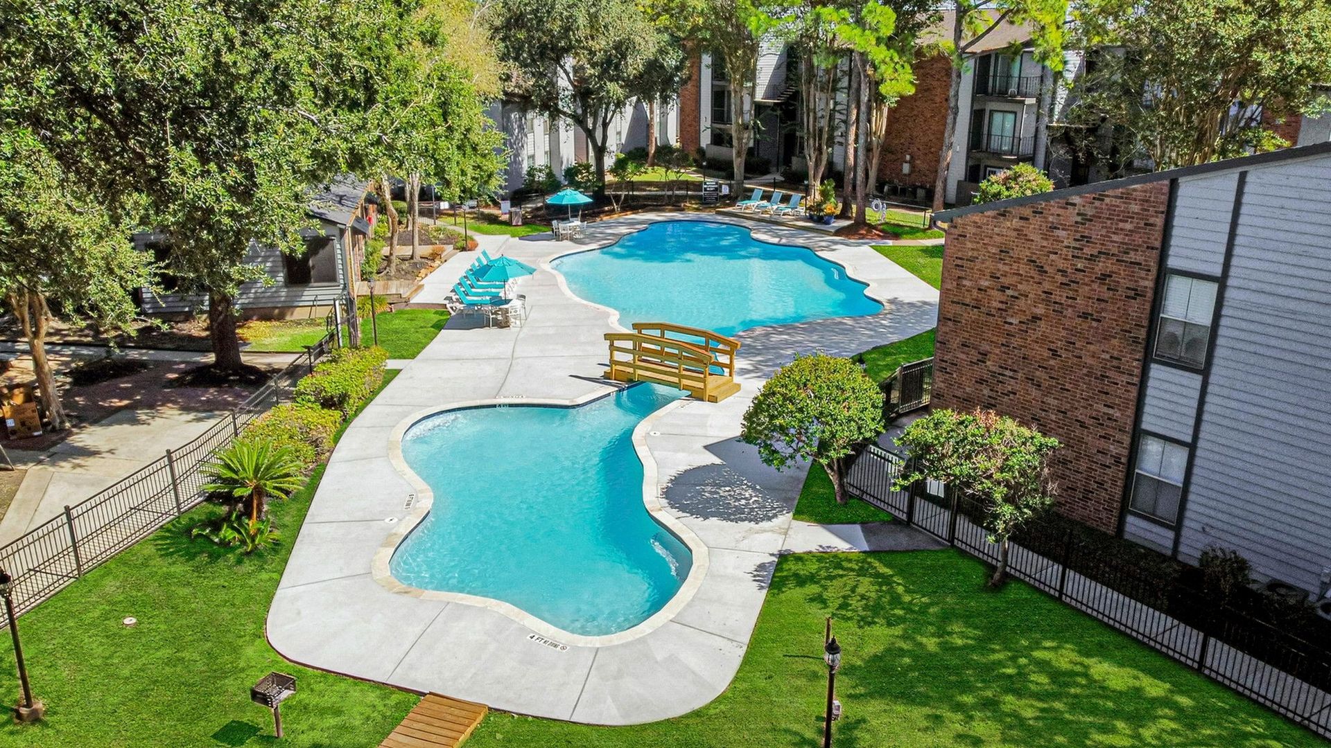Aerial view of a winding outdoor pool in a landscaped apartment community with lounge chairs and a wooden bridge.