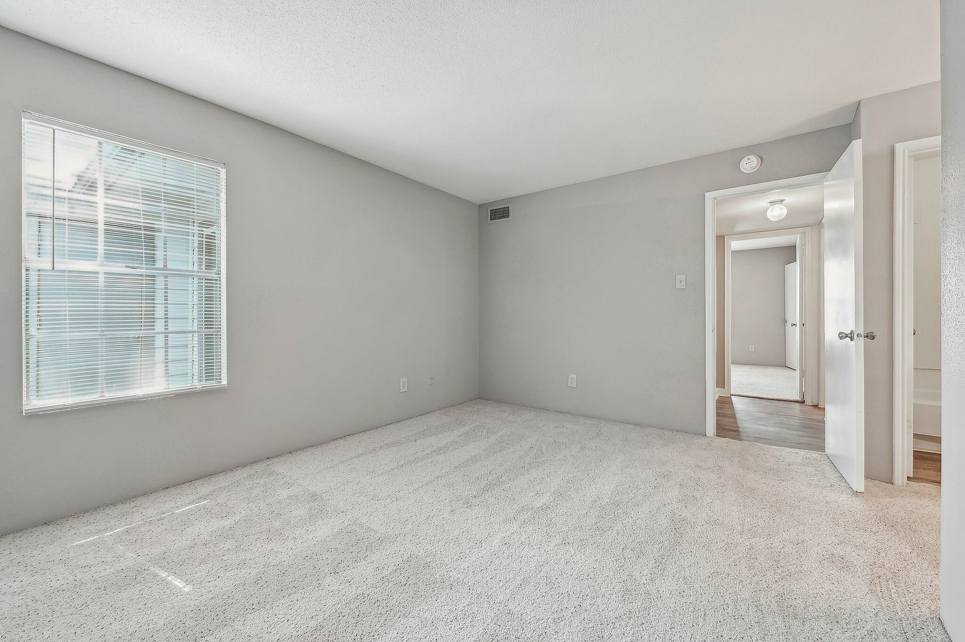 Empty apartment room with window, light carpet, and open doorway.