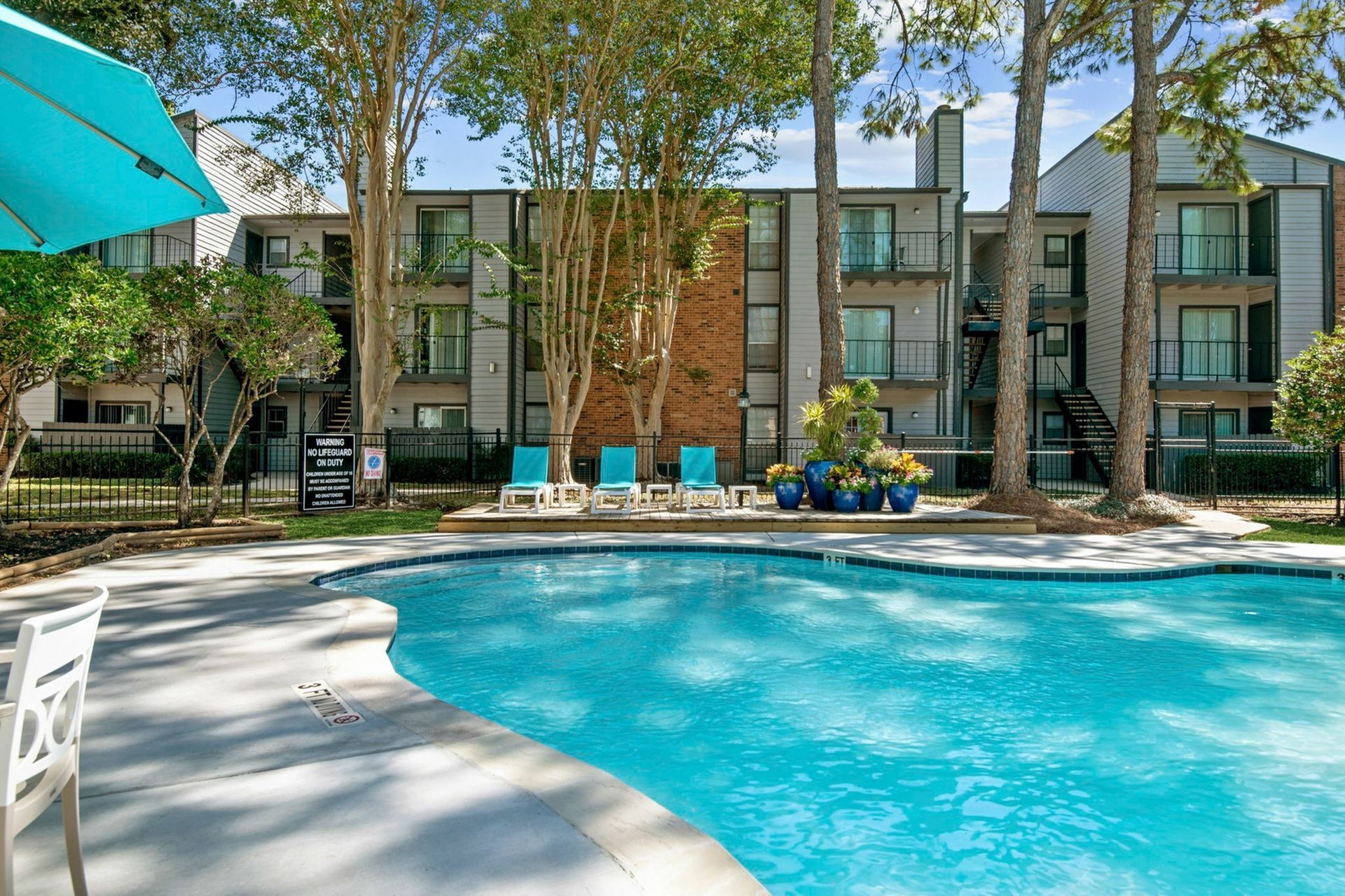 Outdoor apartment community pool with blue lounge chairs, trees, and apartment buildings in the background.