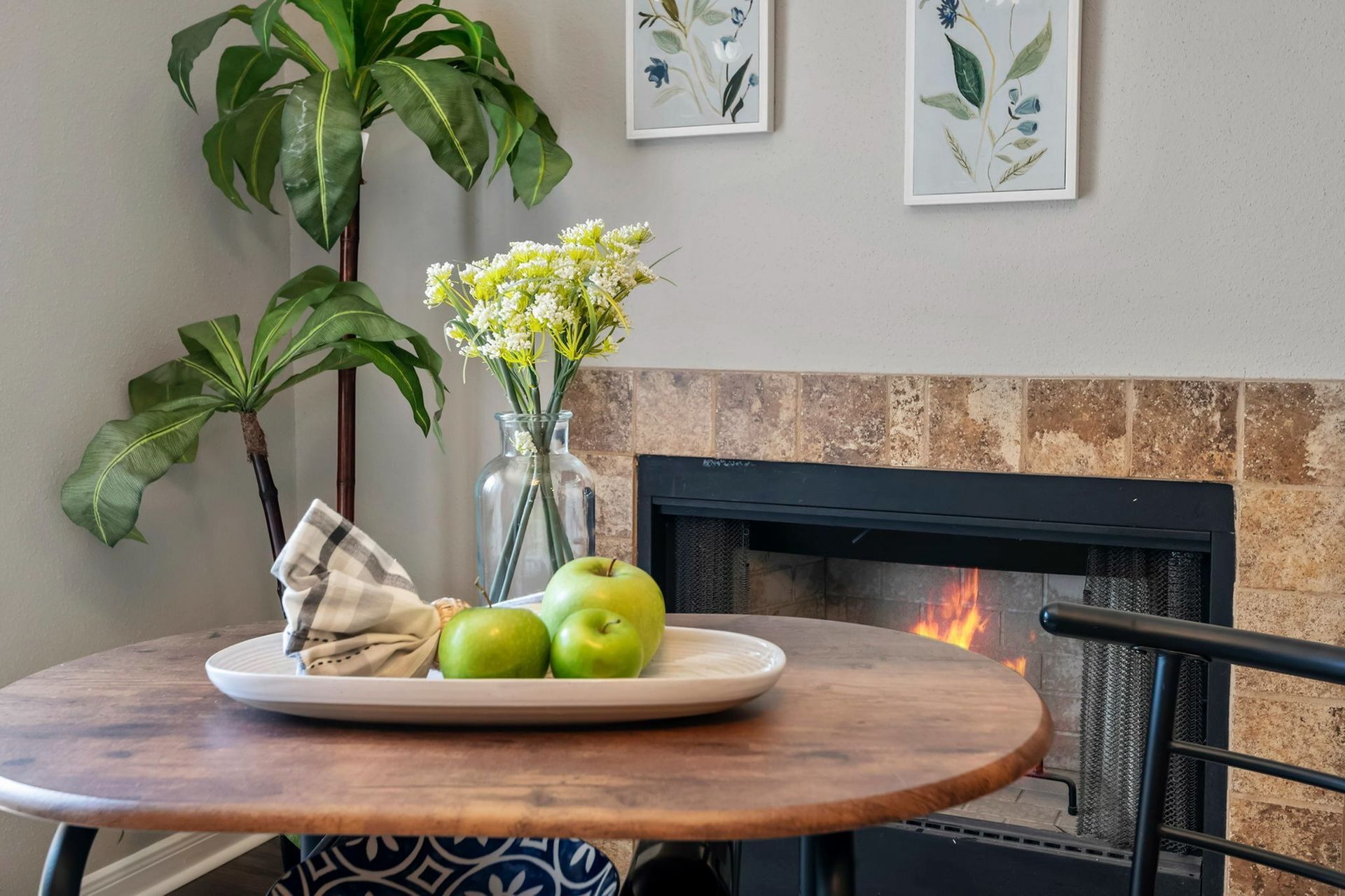 Living room with fireplace, round wooden table, green plant, and a vase of flowers.