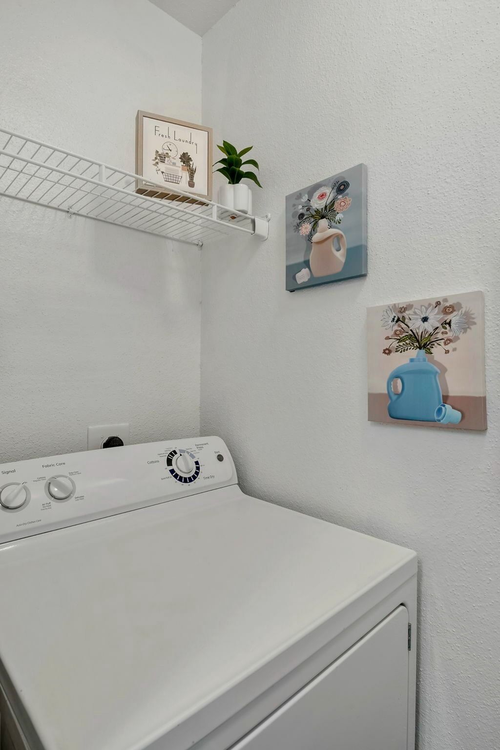 Laundry room with a white top-load washer, wire shelf, and wall art.