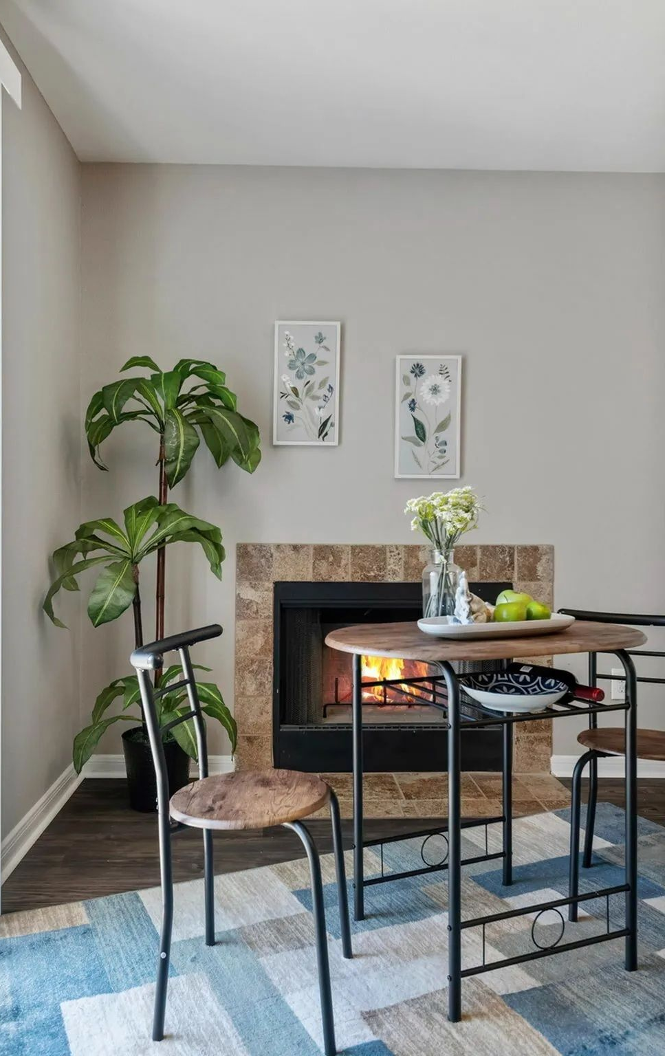 A small dining table with two chairs near a fireplace and a large potted plant in a modern, neutral-toned room.