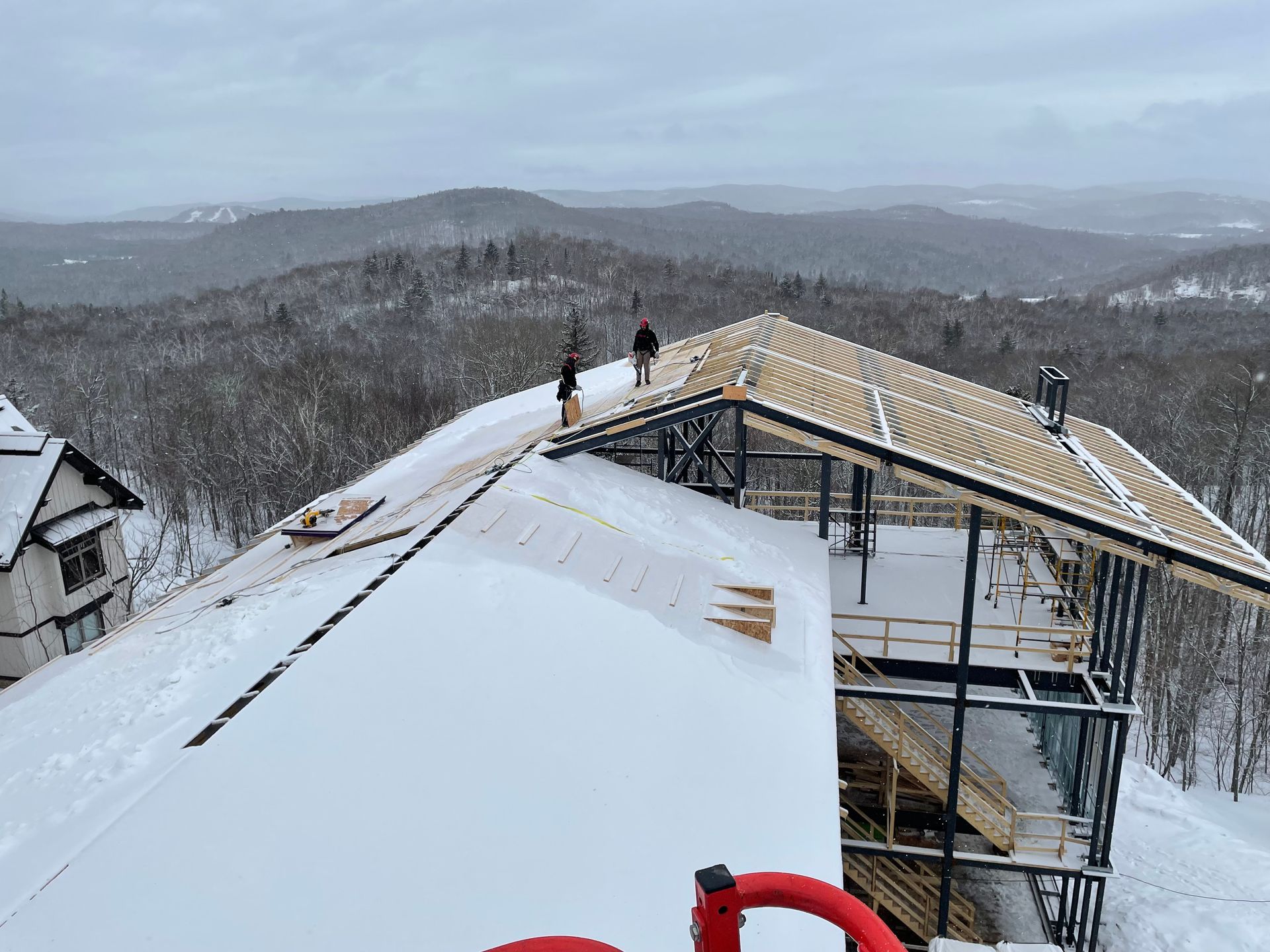 Des ouvriers du bâtiment travaillent sur un toit enneigé, avec les montagnes en arrière-plan. La charpente est terminée, les planches sont posées.