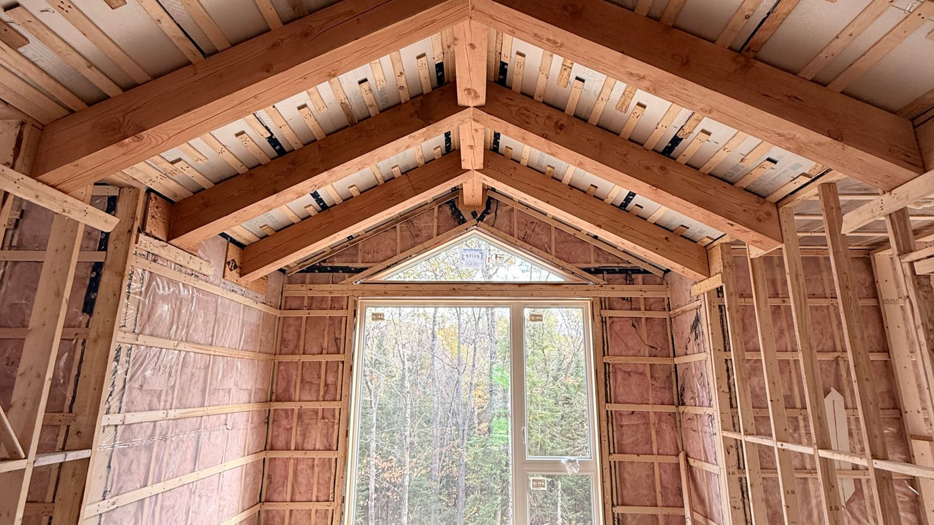 Vue intérieure d'un bâtiment en construction, montrant des poutres en bois, une ossature murale isolée et une fenêtre.