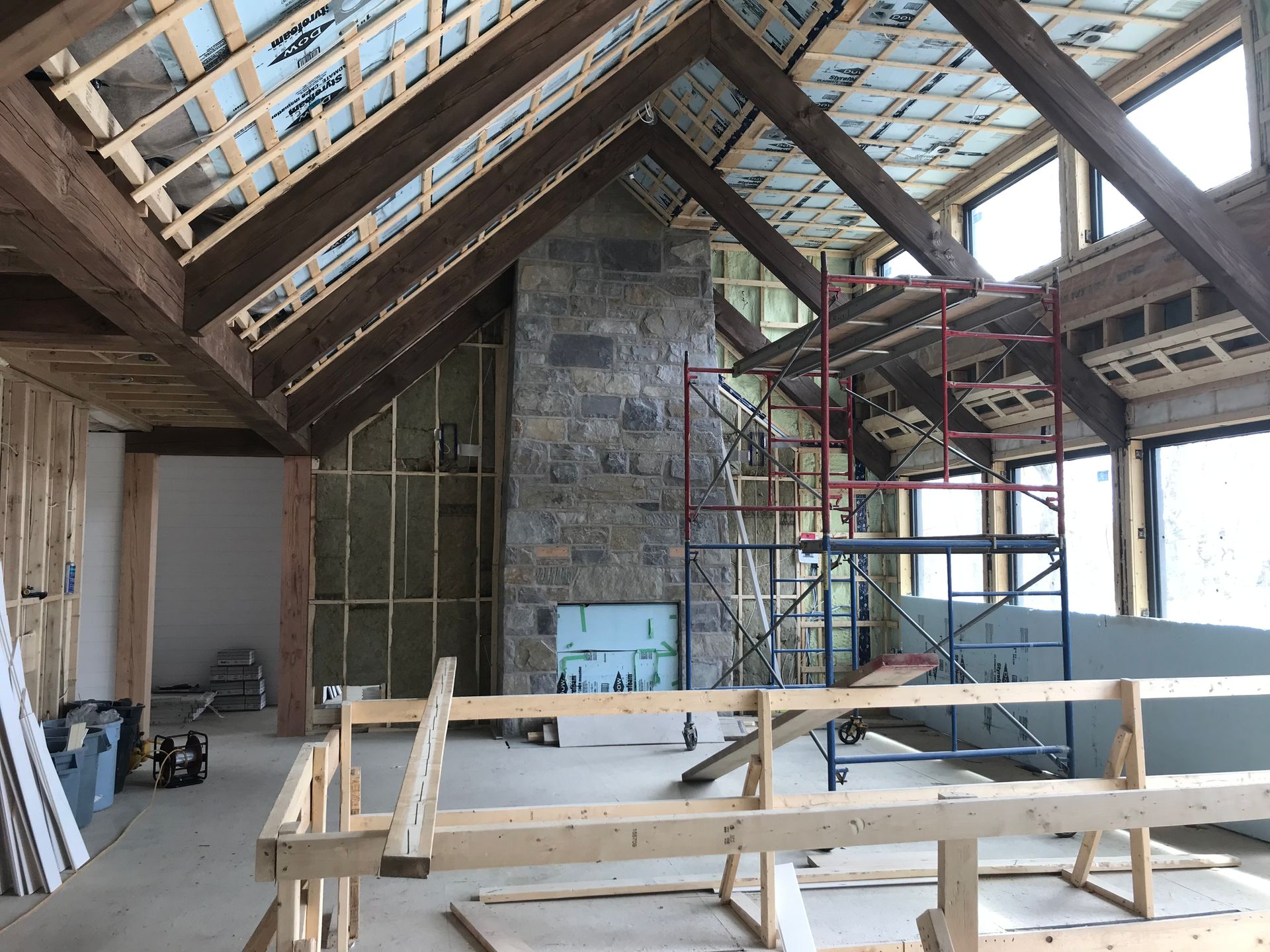 Travaux intérieurs d'une maison. Poutres en bois apparentes et cheminée en pierre ; échafaudages et isolation visibles.