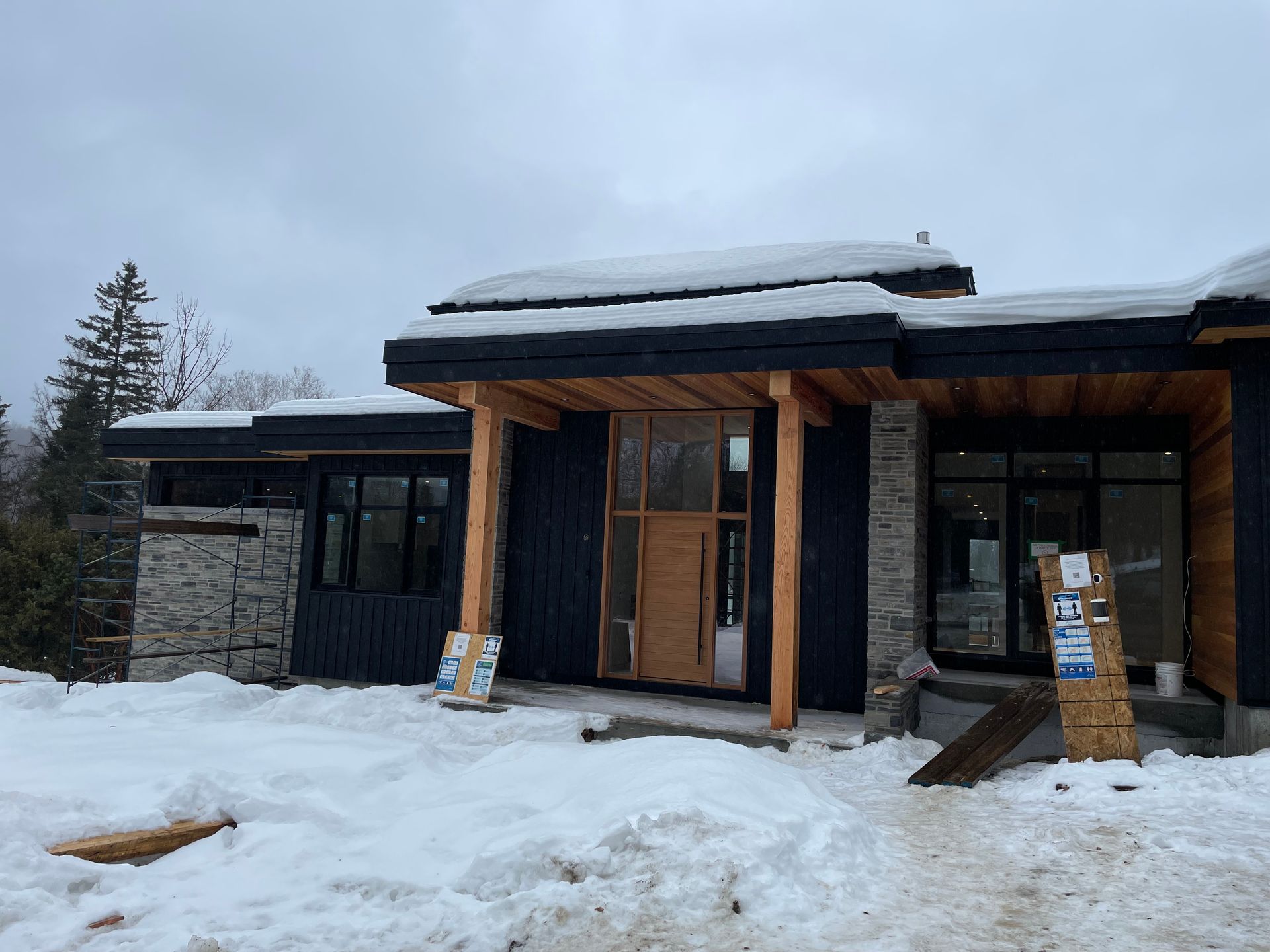 Une maison moderne en construction, bardage noir, éléments en pierre, neige au sol et sur le toit, ciel nuageux.