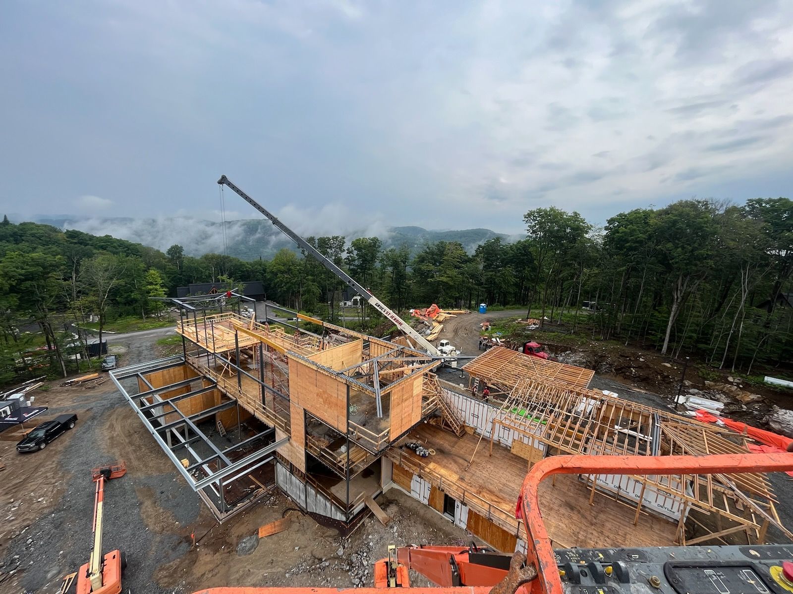 Chantier de construction avec une maison en construction, une grue et des arbres sous un ciel nuageux.