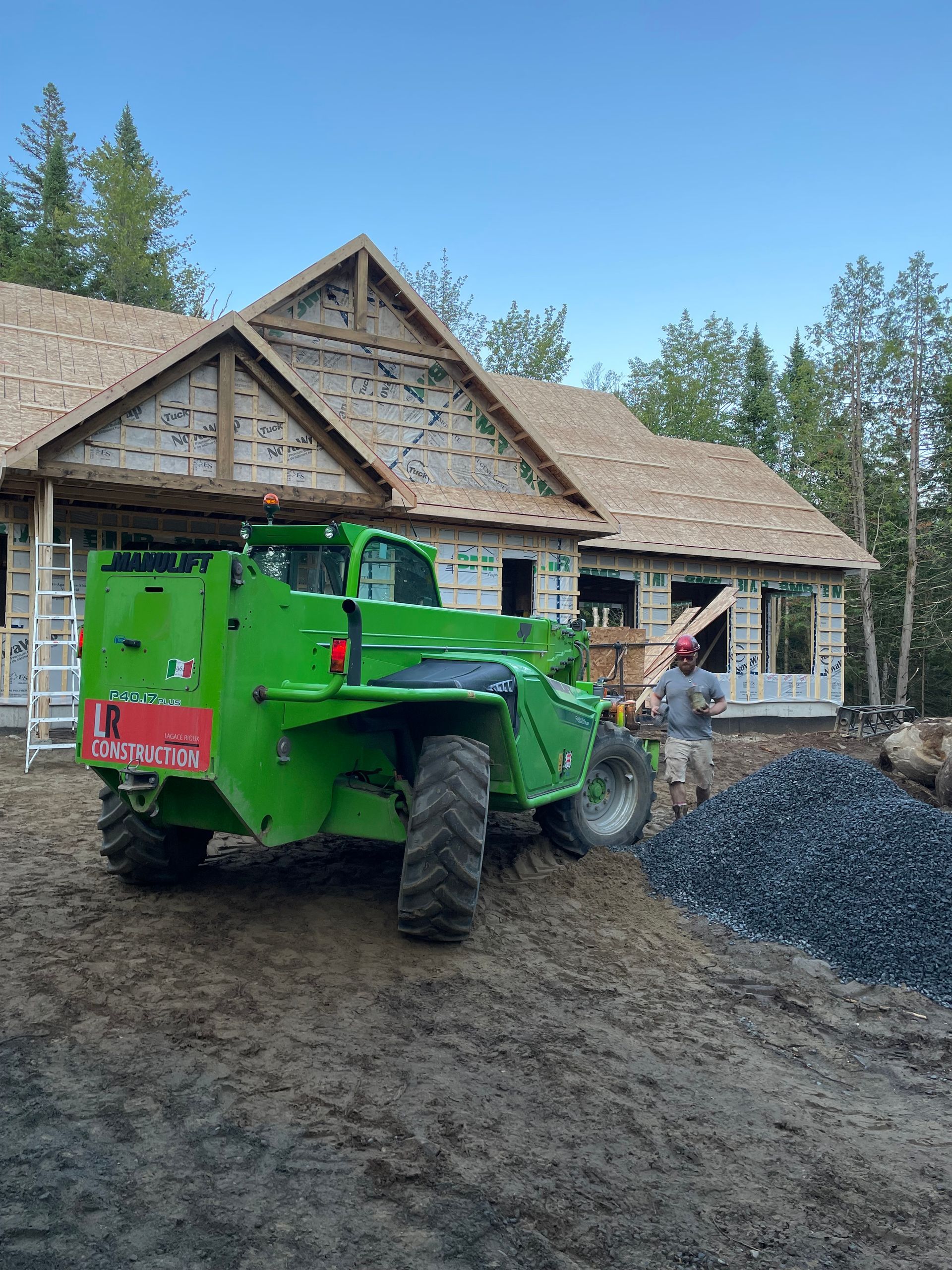 Un chariot élévateur de chantier vert se trouve près d'une maison en construction. Un ouvrier se tient à proximité avec du gravier.