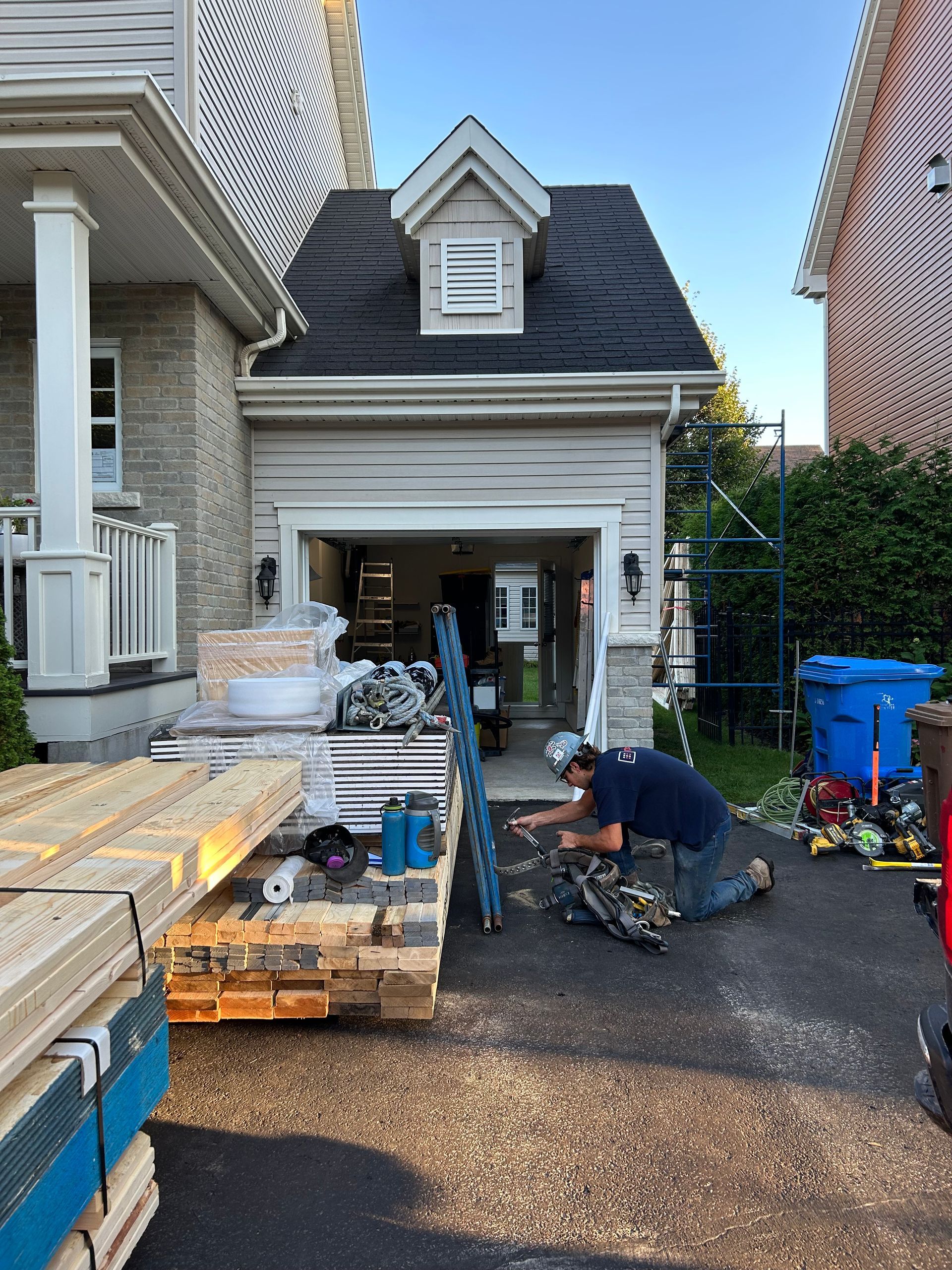 Une personne est agenouillée sur une allée devant un garage, en train de travailler sur des matériaux de construction. Du bois et des outils sont à proximité.