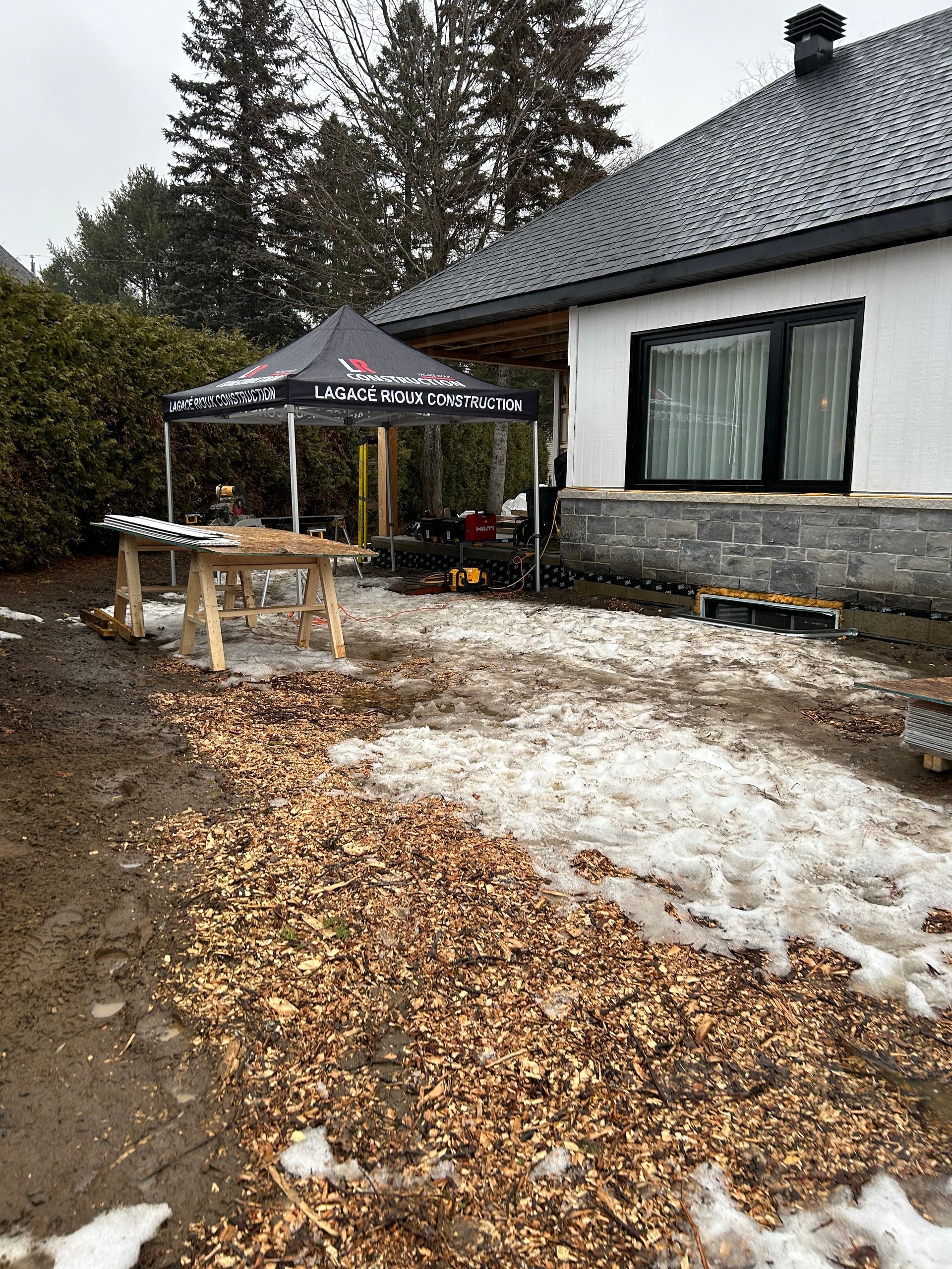 Zone de travail extérieure avec copeaux de bois, une tente et un abri. Conditions enneigées et boueuses.