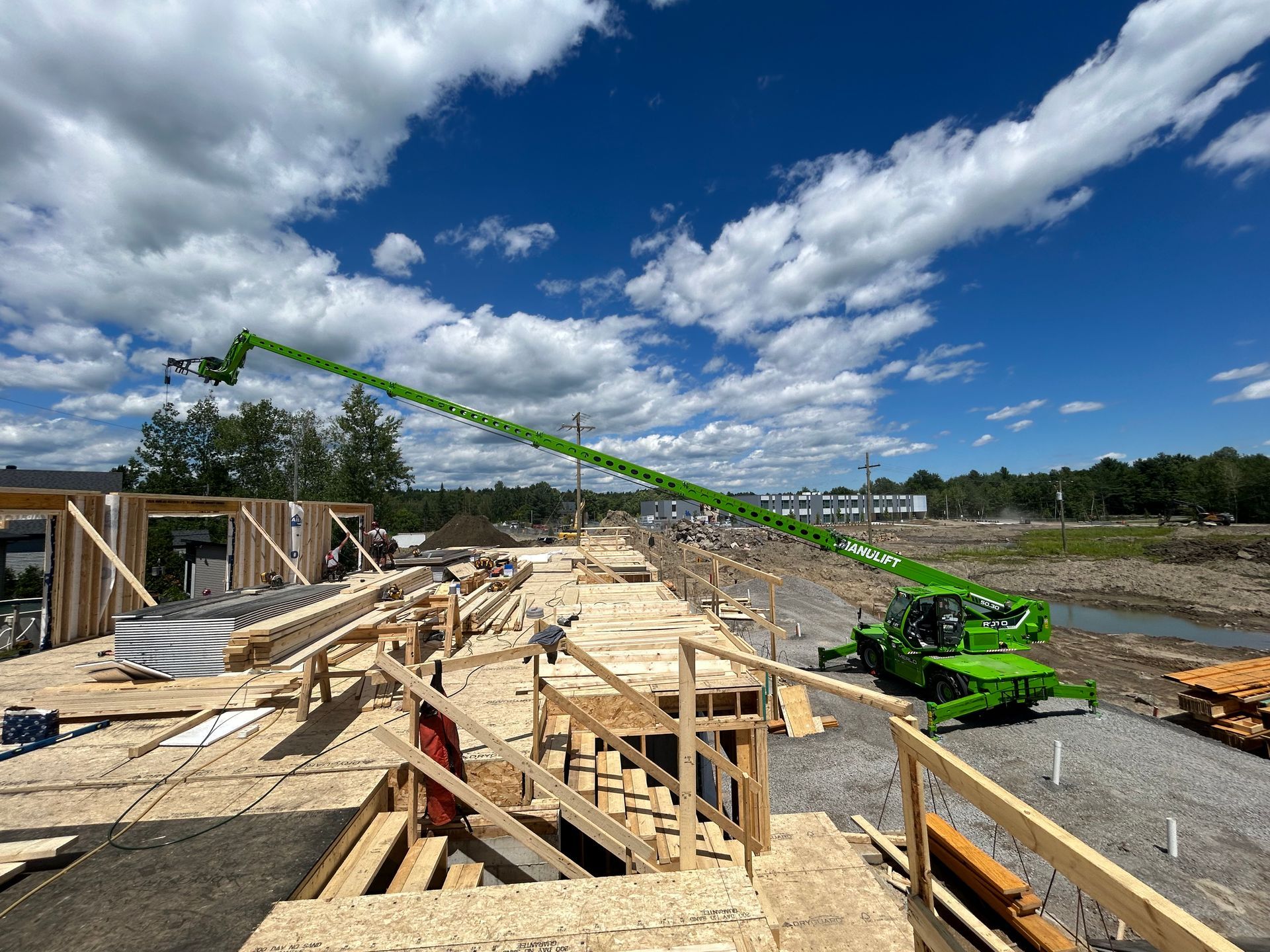 Chantier de construction avec une longue nacelle élévatrice verte déployée sur un ciel bleu, des structures en bois et un bâtiment partiellement achevé.
