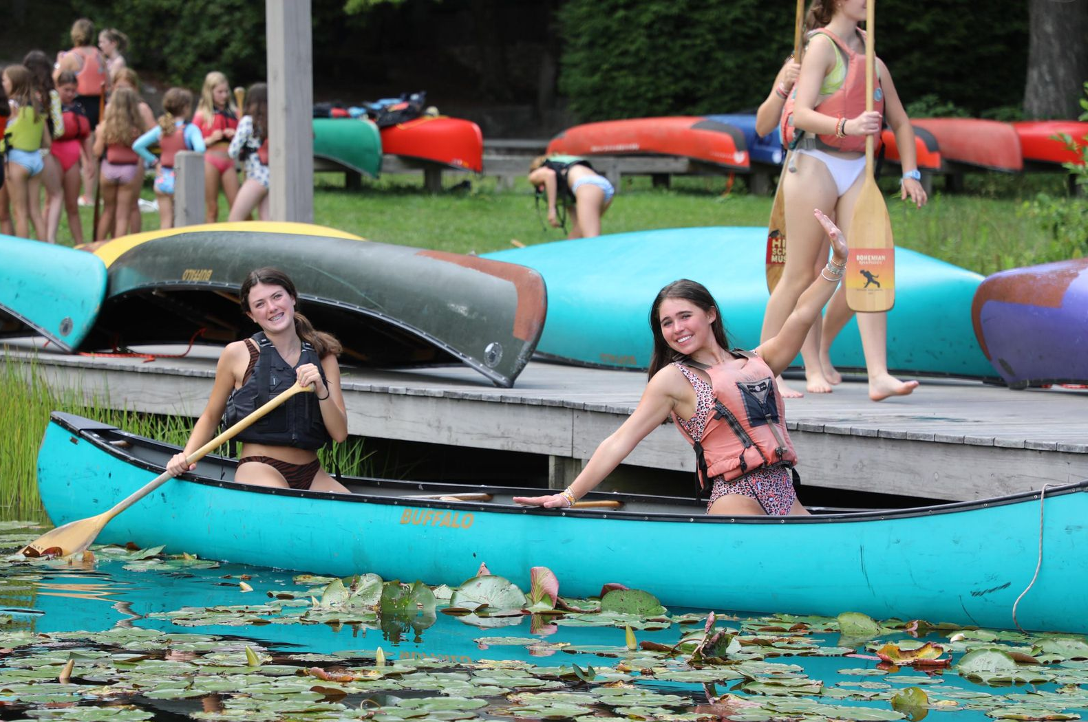 Two people smile in a teal canoe on a lily-pad-covered lake near a wooden dock with other canoes and people.