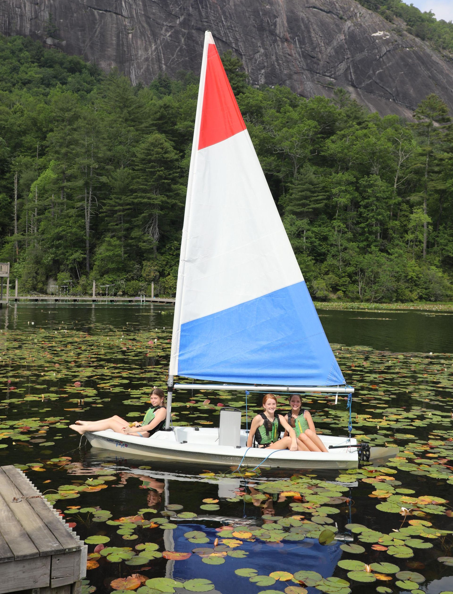 Three people on a small sailboat with a red, white, and blue sail on a lake filled with lily pads near a rocky cliff.