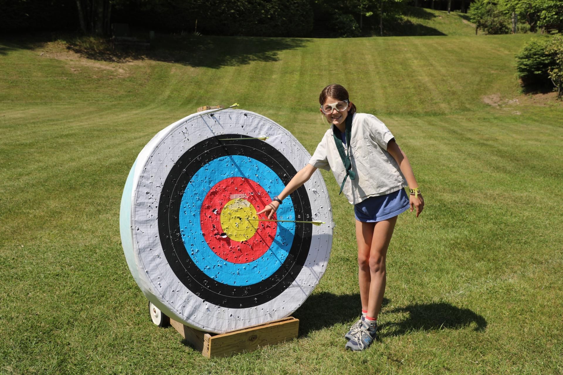 A person points to an arrow hitting the center of a large, circular archery target on a grassy field.