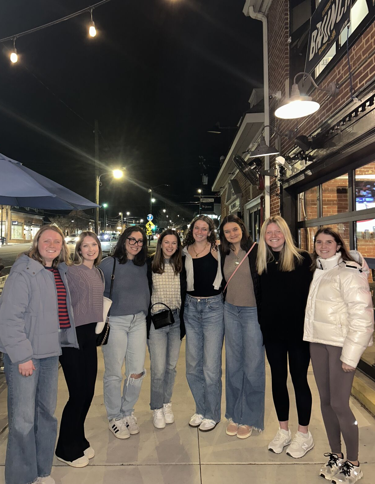 Eight people stand together in a row smiling for a photo outside a lit storefront at night.