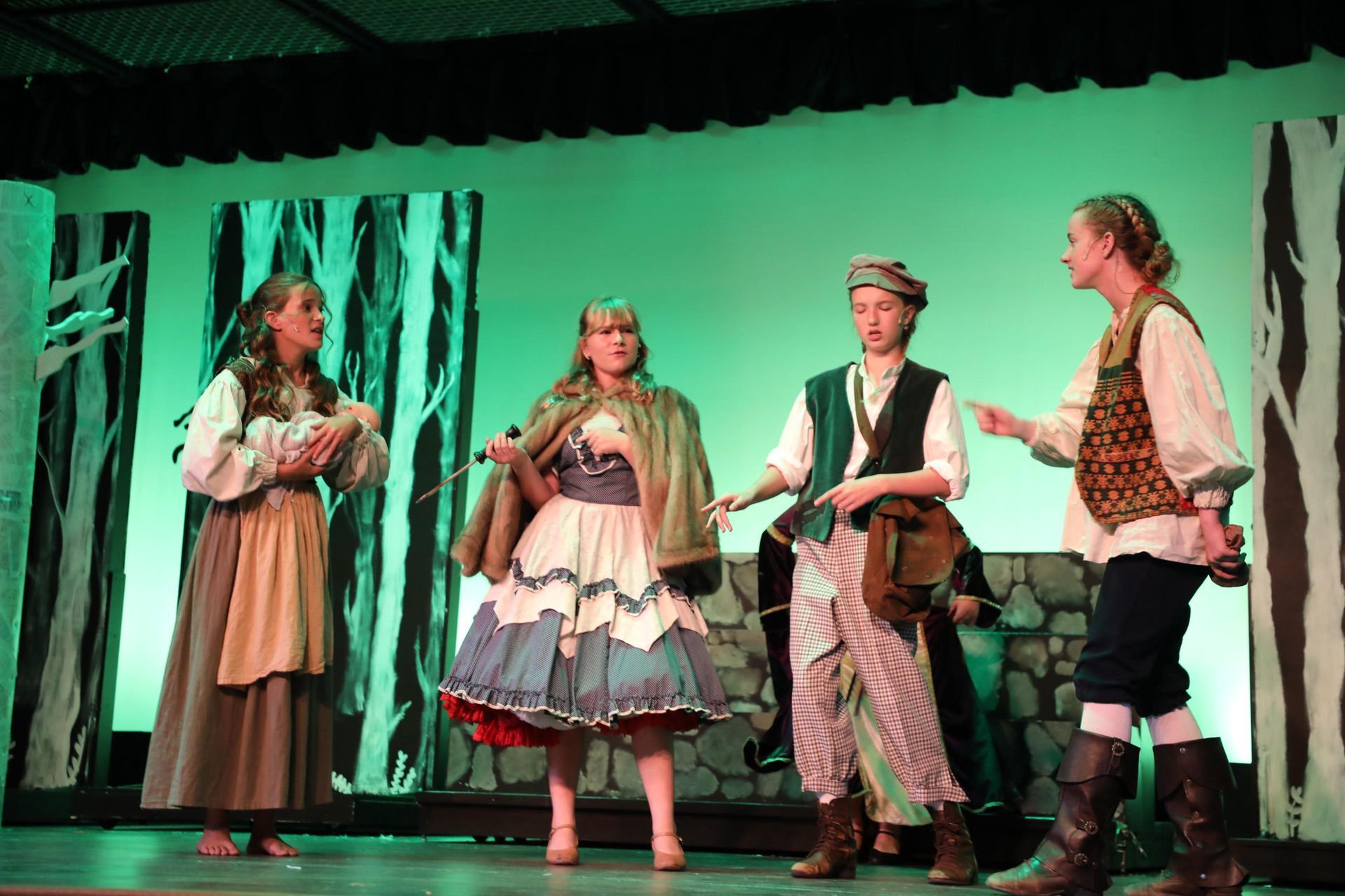 Four actors in historic-style costumes perform a scene on a stage with a forest backdrop.