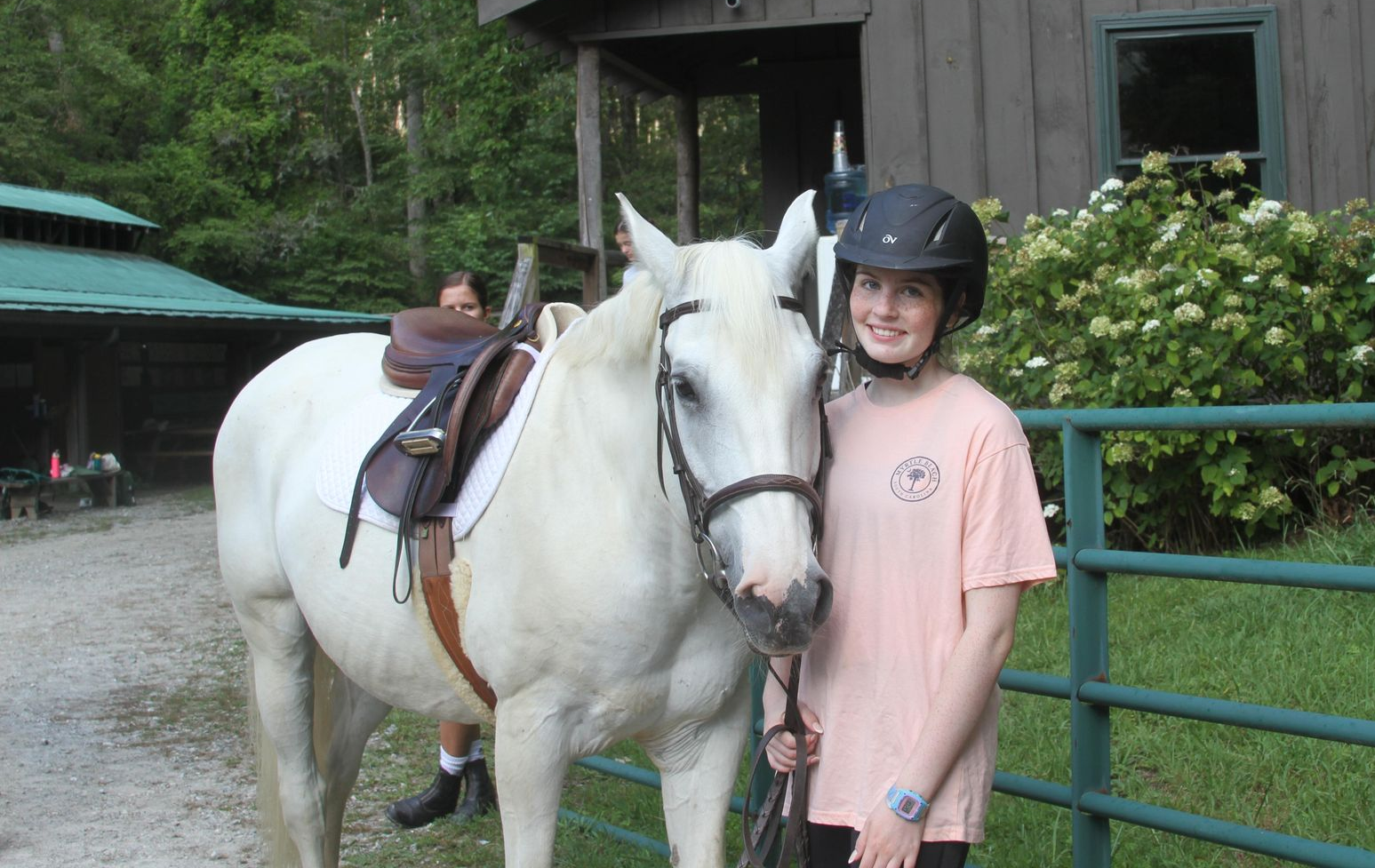A person wearing a helmet and peach shirt stands beside a saddled white horse near a wooden building and green fence.