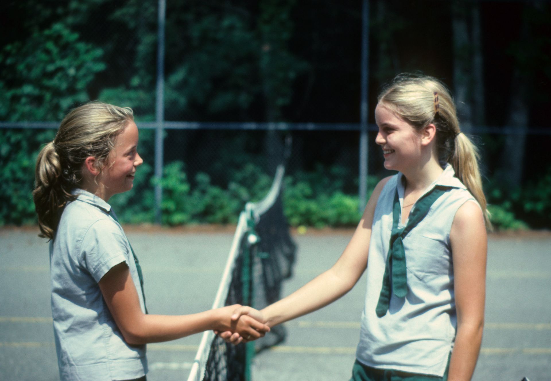 Two people in matching gray uniforms shake hands on a tennis court, smiling at each other.