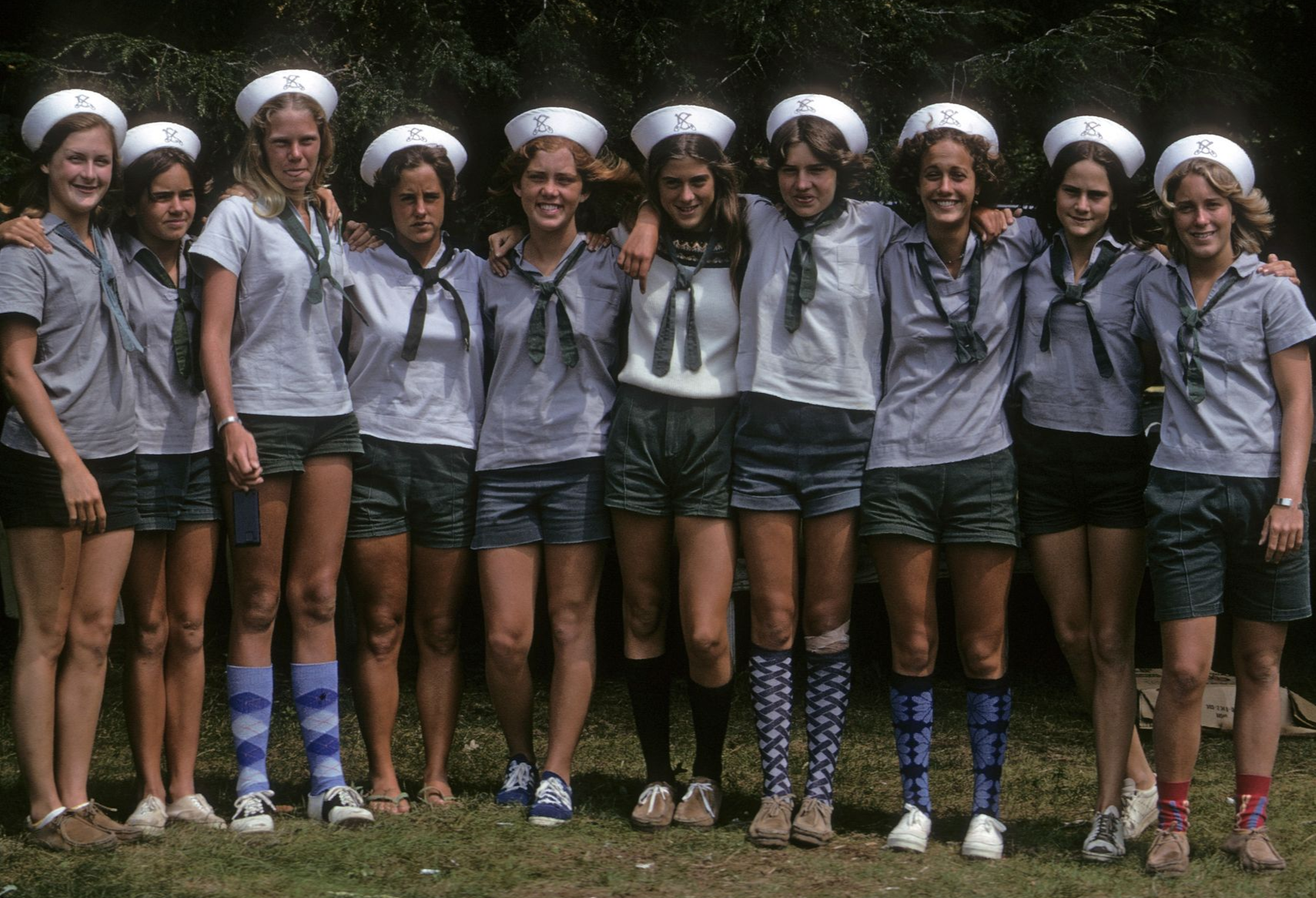 A group of eleven people in matching sailor-style uniforms with hats, grey shirts, and dark shorts, posing outdoors.