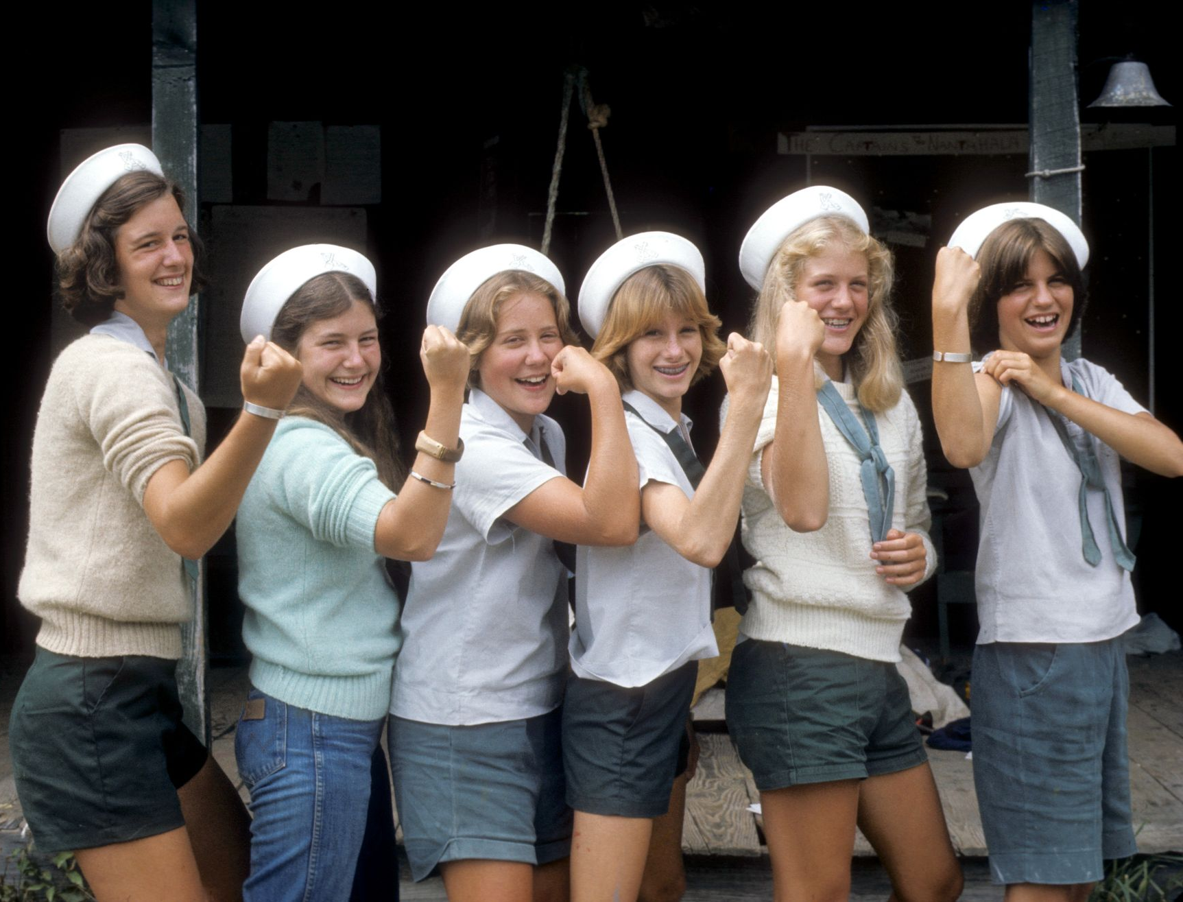Six people wearing white sailor hats and shorts stand in a row, smiling and flexing their arms for the camera.