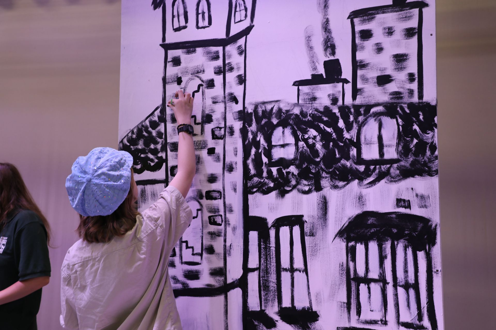 A person wearing a light blue cap paints a large black-and-white mural of a multi-story building on a wall.