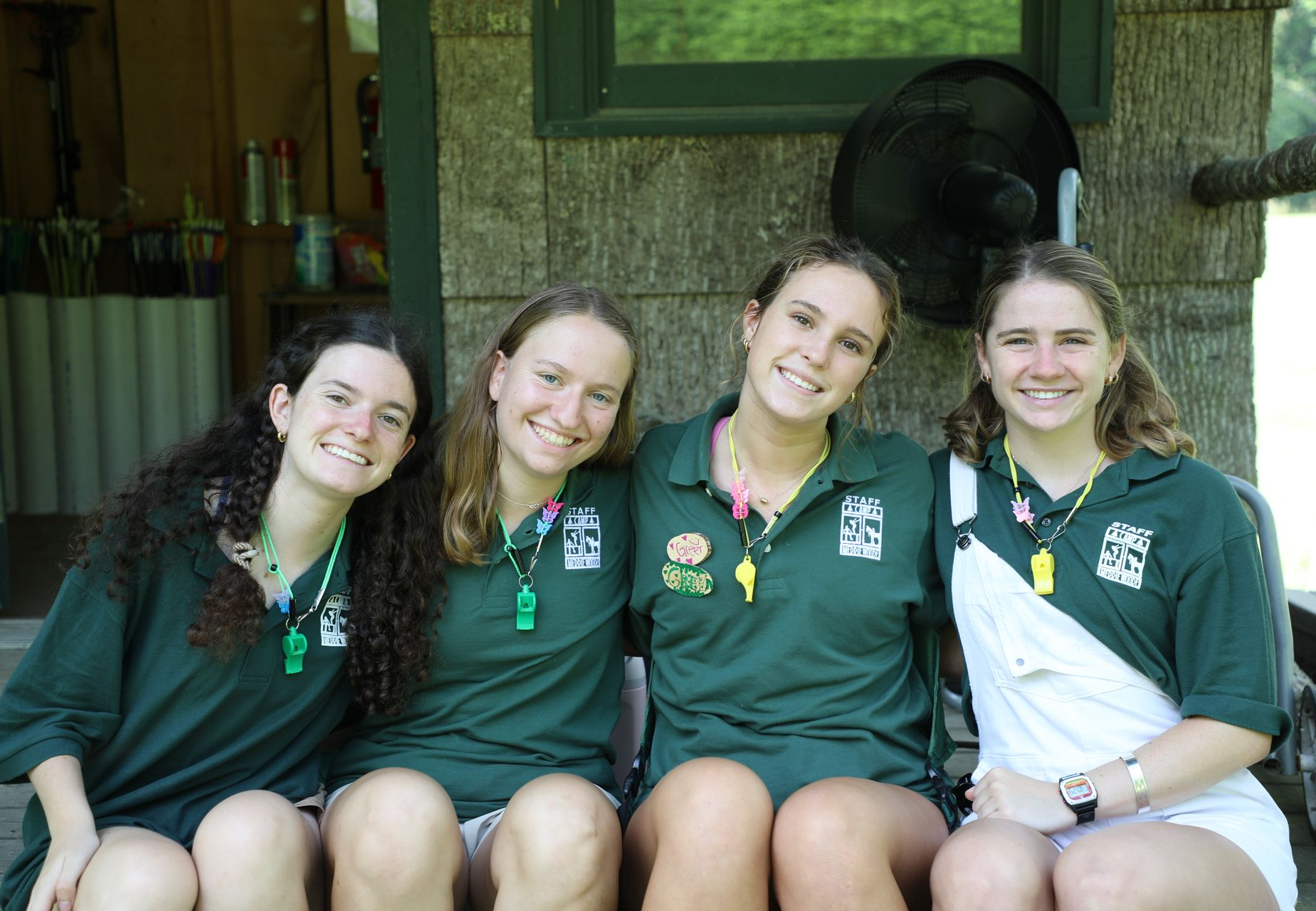 Four smiling people in matching green polo shirts and whistles sit together outdoors in front of a wood-paneled building.