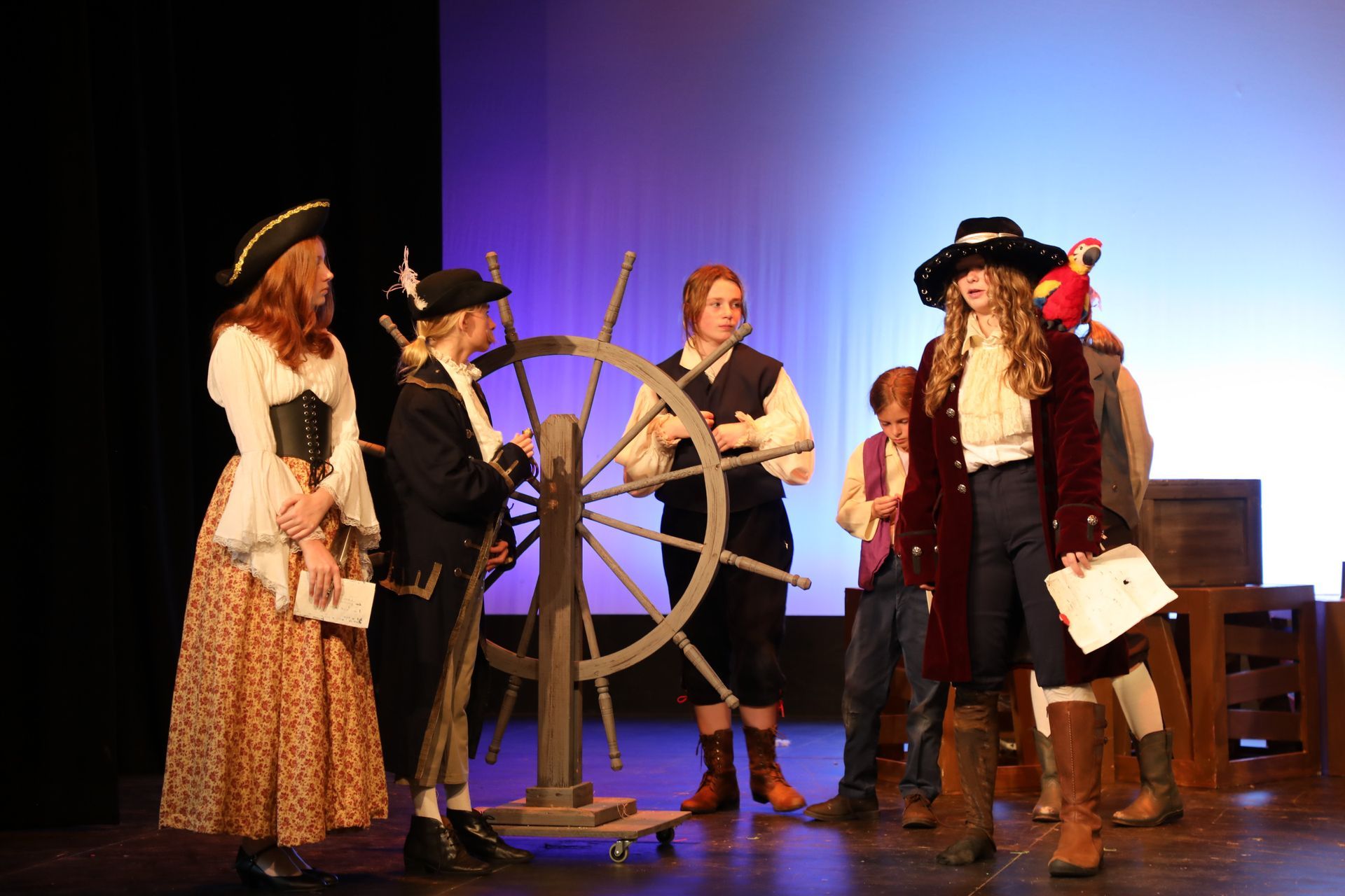 Actors in historical pirate costumes stand on a stage around a wooden ship's wheel.