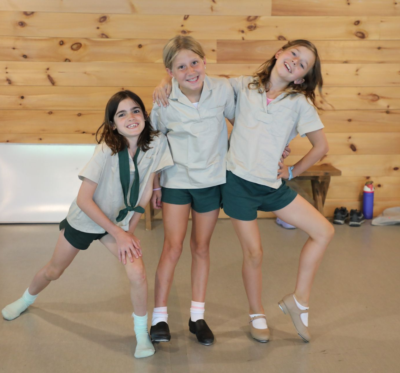 Three people in matching tan shirts and dark green shorts pose playfully in front of a wood-paneled wall.
