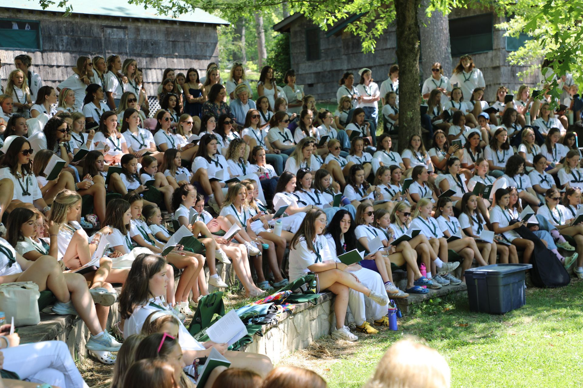 A large group of people in white attire sits on stone steps and grass, gathered for an outdoor event under trees.