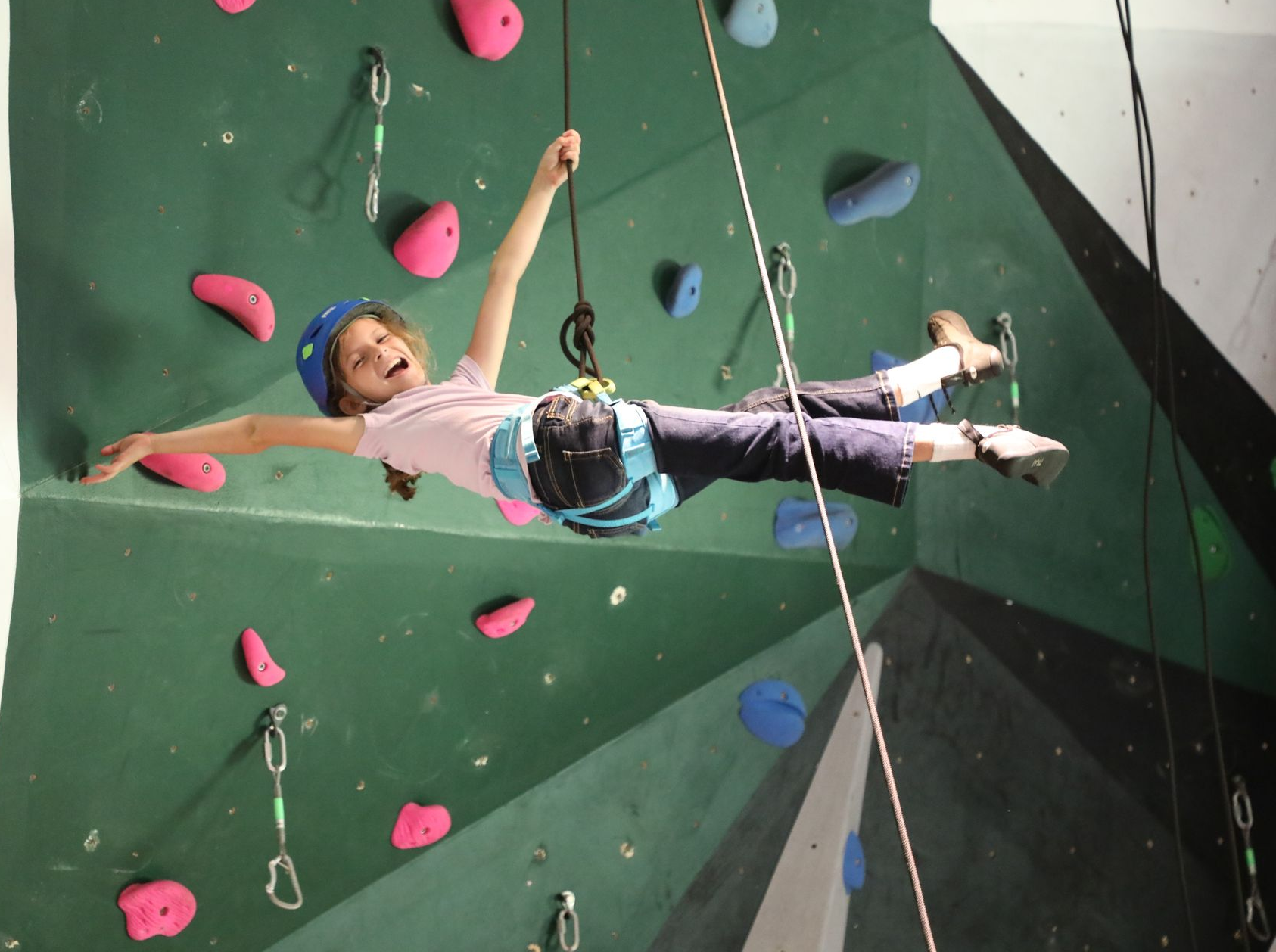A person in a climbing harness joyfully jumps mid-air against a green indoor climbing wall with colorful holds.