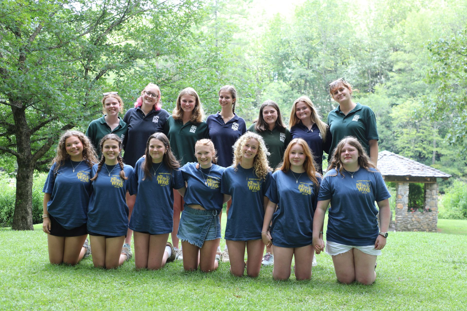 A group of 14 people smiling and posing in two rows on a grassy lawn in front of trees and a stone gazebo.