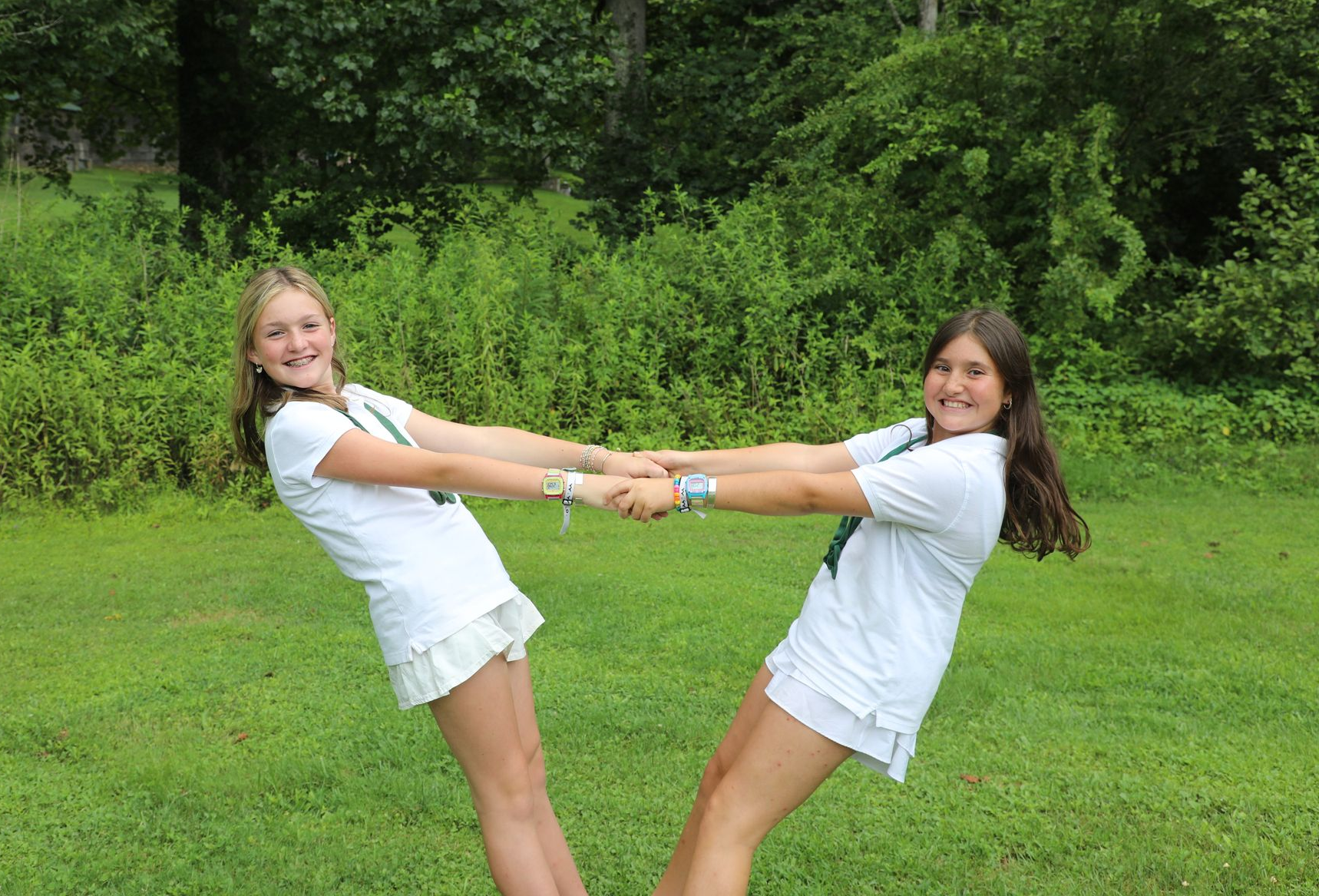 Two people in white shirts and shorts hold hands and lean backward in a grassy park, smiling at the camera.