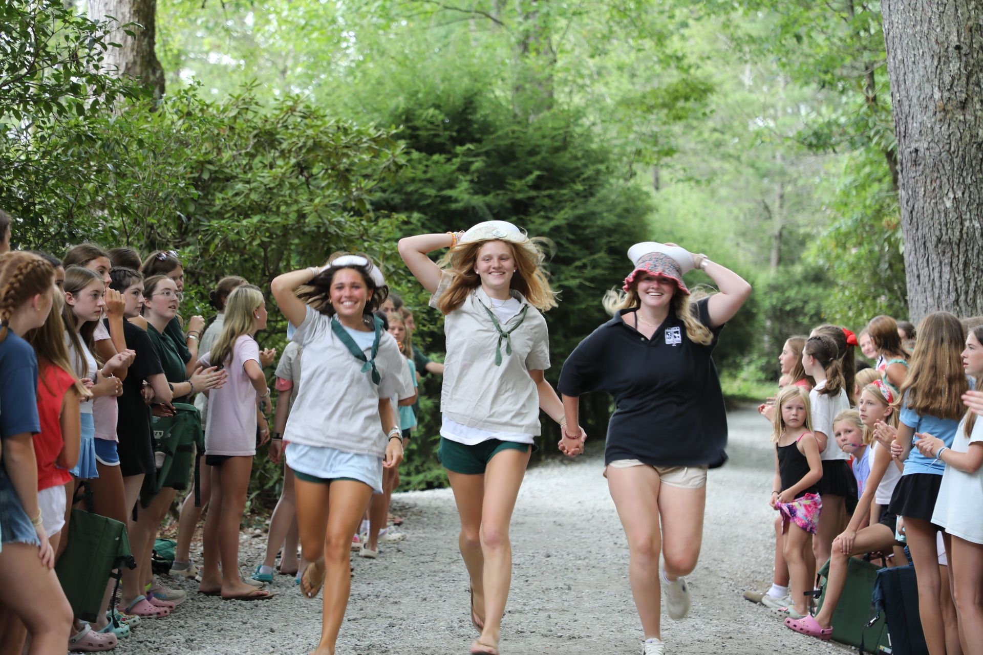 Three people running on a gravel path through a wooded area, holding hats on their heads while others watch from sides.