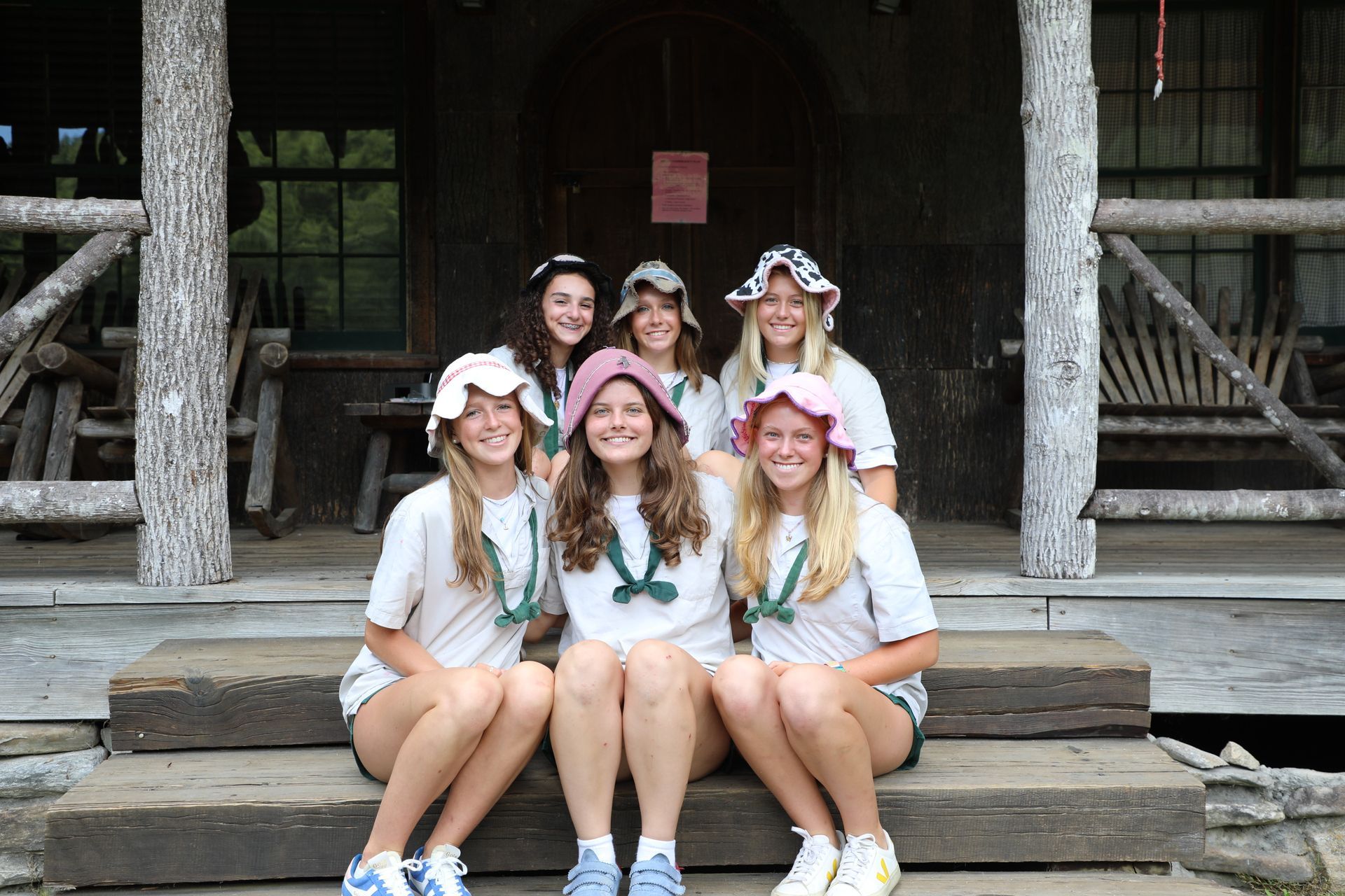 Six young people in matching white shirts and bucket hats sit together on the steps of a wooden cabin.