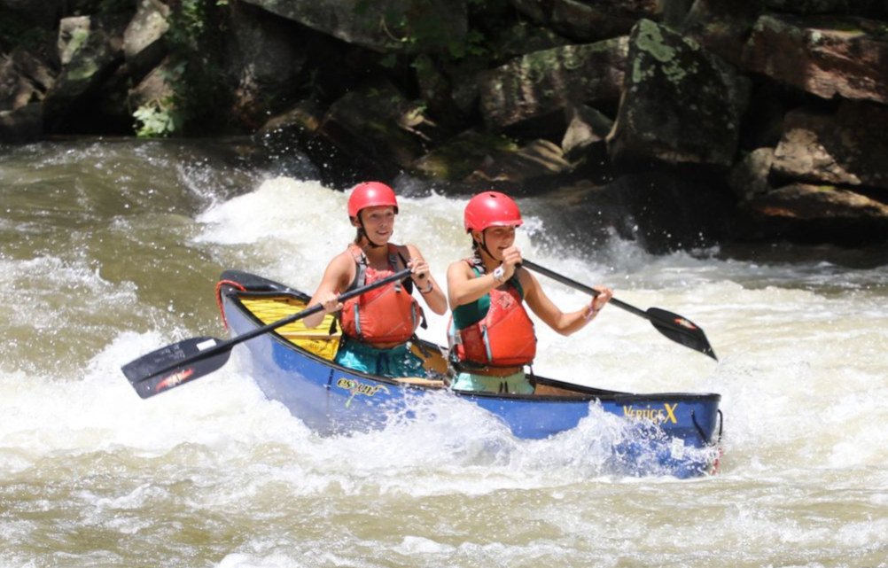 Two people wearing red helmets and life vests paddle a blue canoe through white-water rapids next to rocky shores.