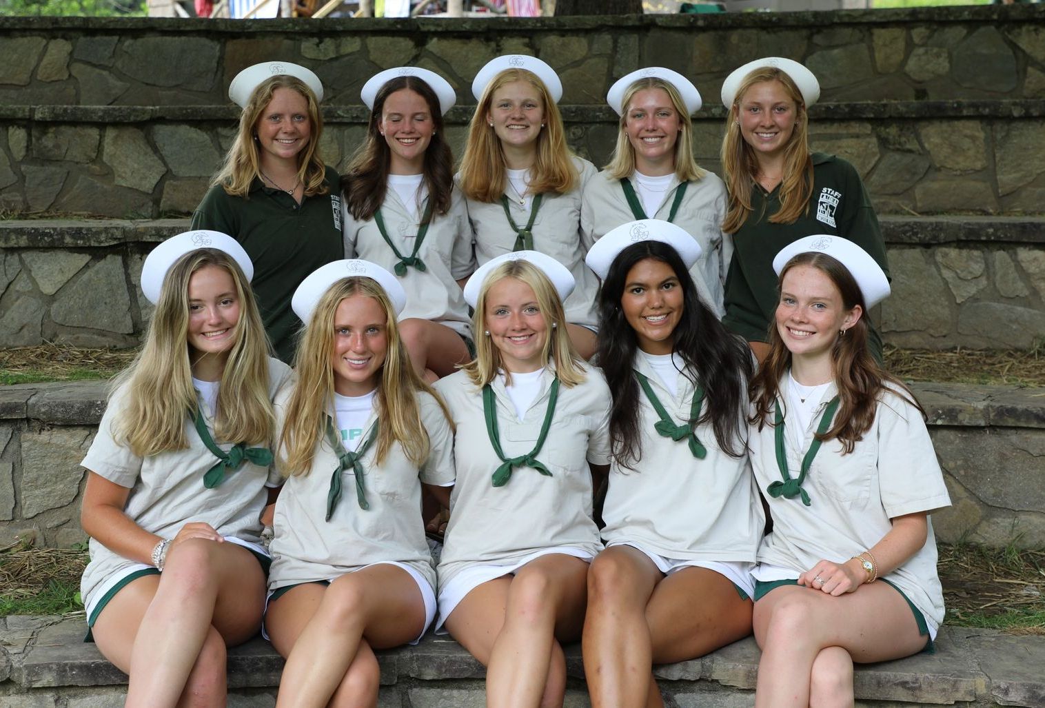 Ten individuals wearing white berets and scarves sit in two rows on stone steps outdoors, smiling at the camera.