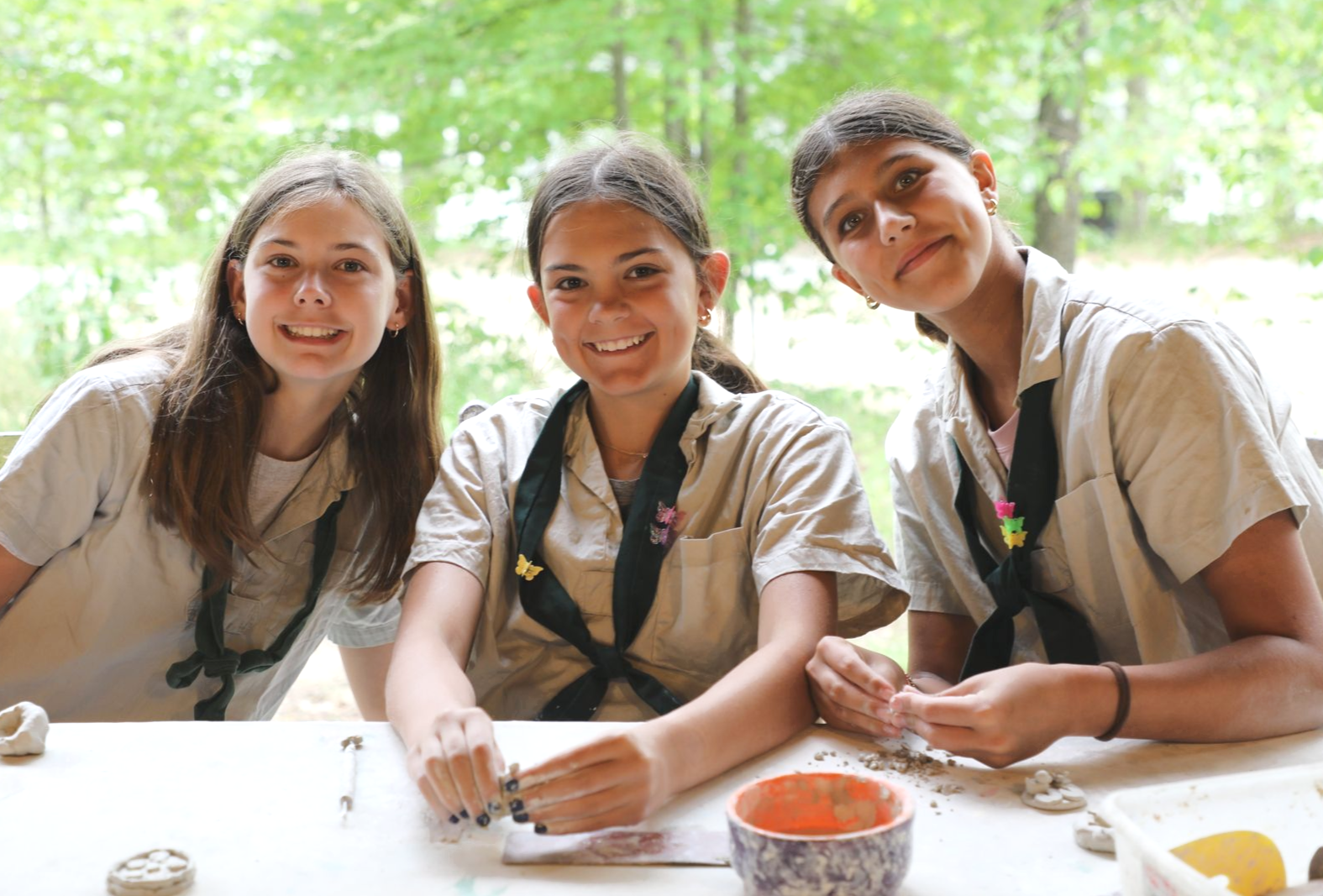 Three smiling young people in uniform scarves work with clay at a table in a wooded setting.