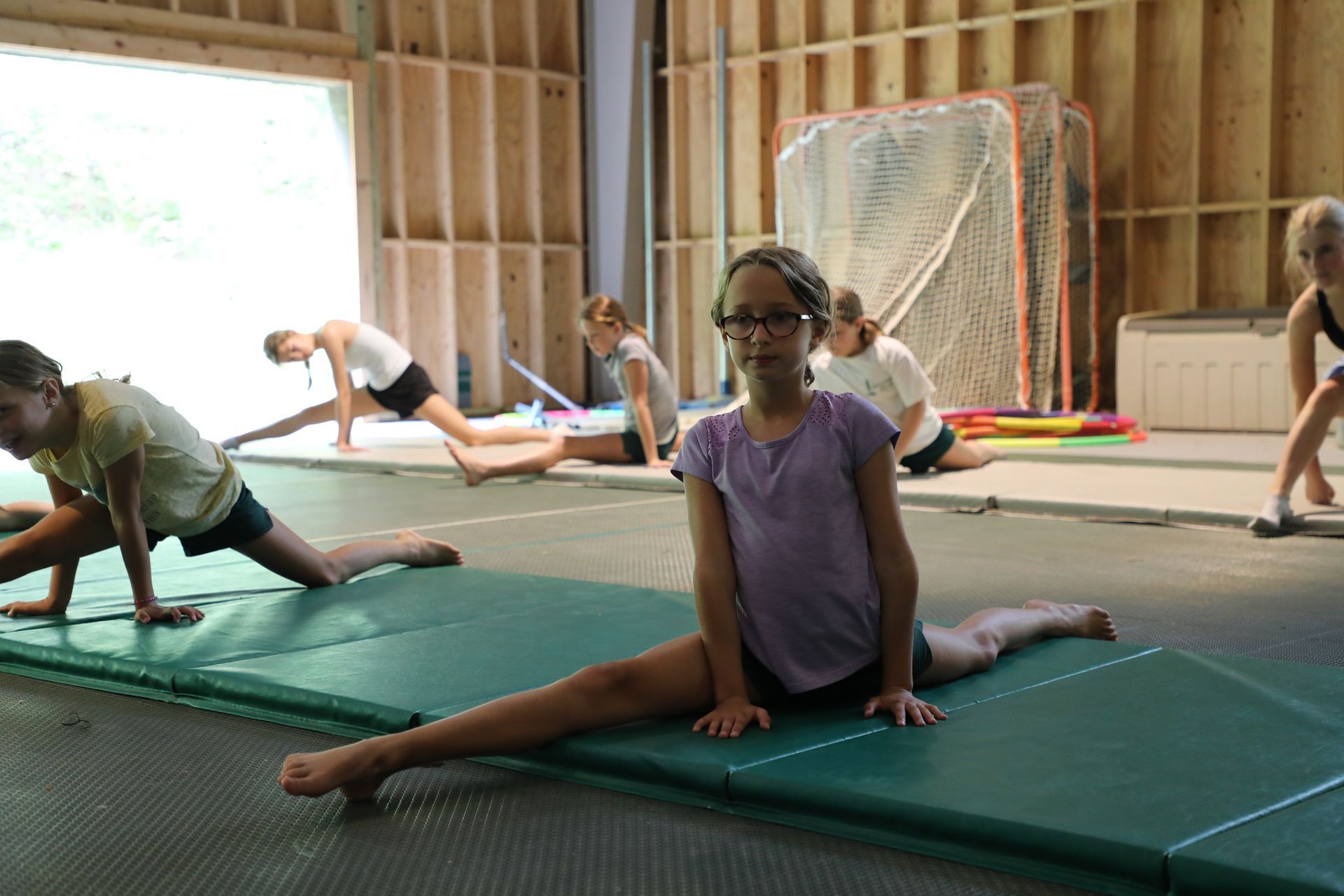 A group of people practices floor splits on green mats inside an unfinished wooden building.